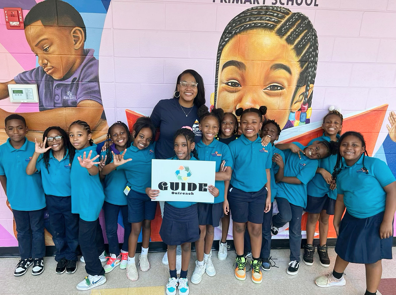 Group of children in blue uniforms holding a guide sign, standing in front of a colorful mural of a smiling face at school.