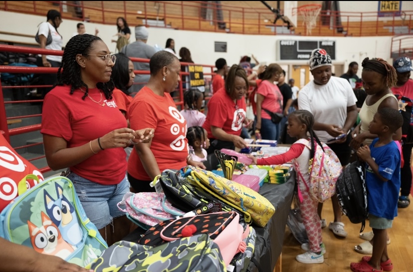 Group of people, including children and adults, gathered around tables with backpacks and supplies in a gymnasium.