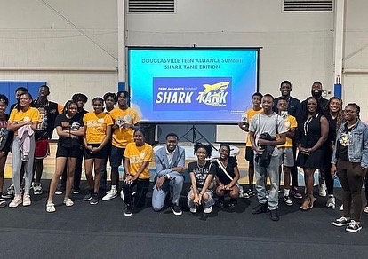 Group of people in yellow and black shirts standing and sitting in front of a large screen displaying 'Shark Tank' at an event.