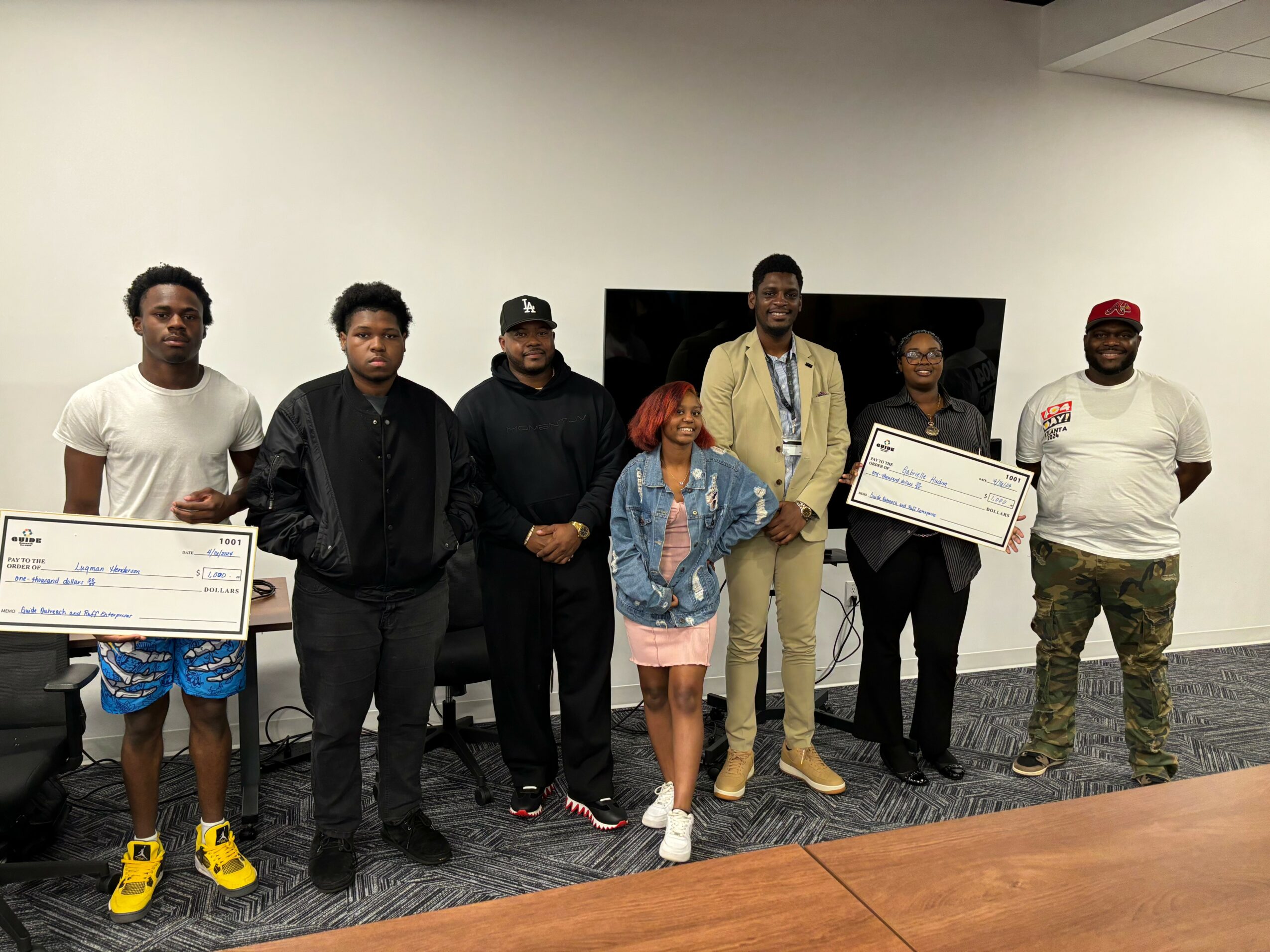 Group of seven people standing indoors, some holding large checks, with a TV screen behind them.