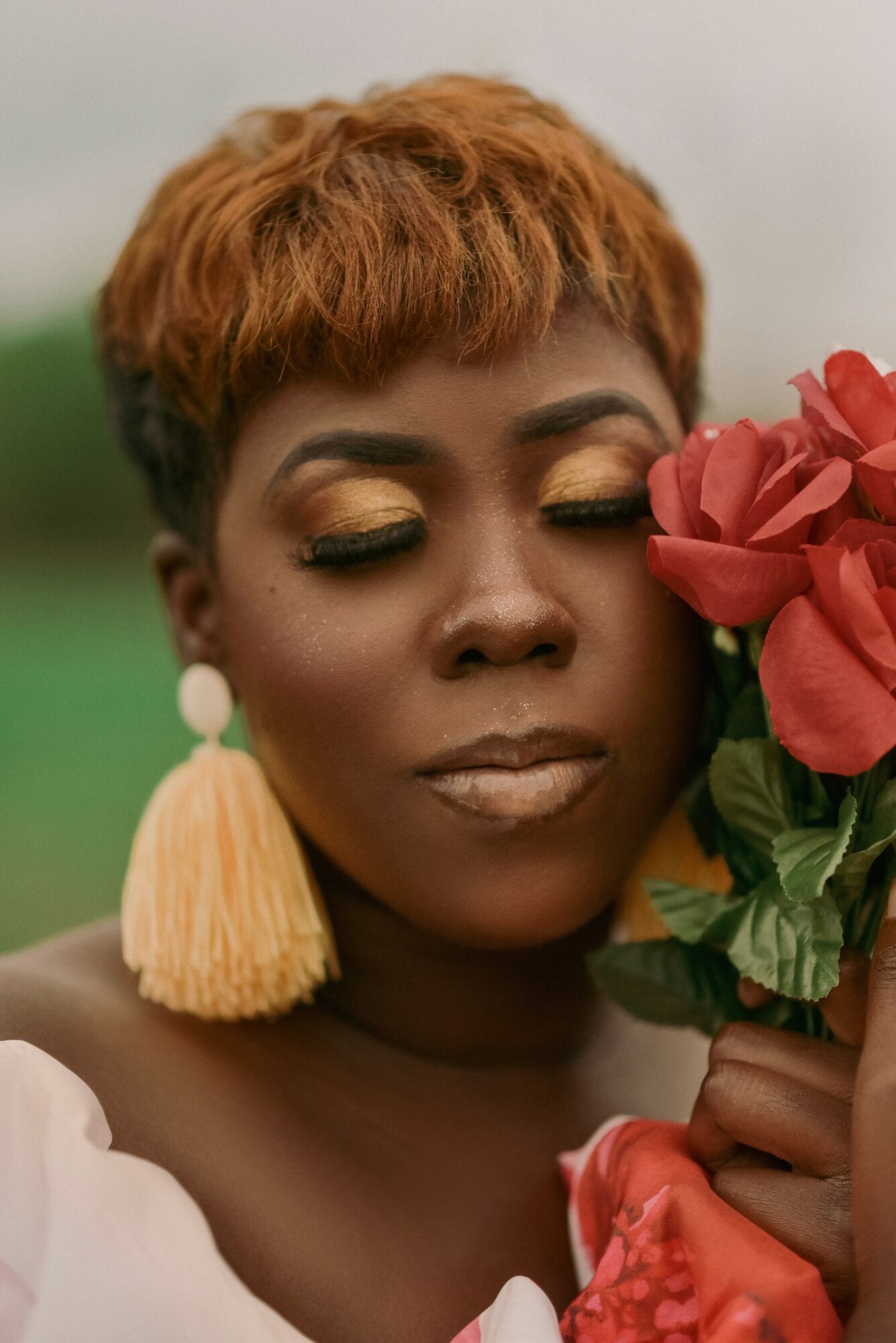 Woman with short reddish hair and gold eye makeup holding pink flowers, wearing large tassel earrings.