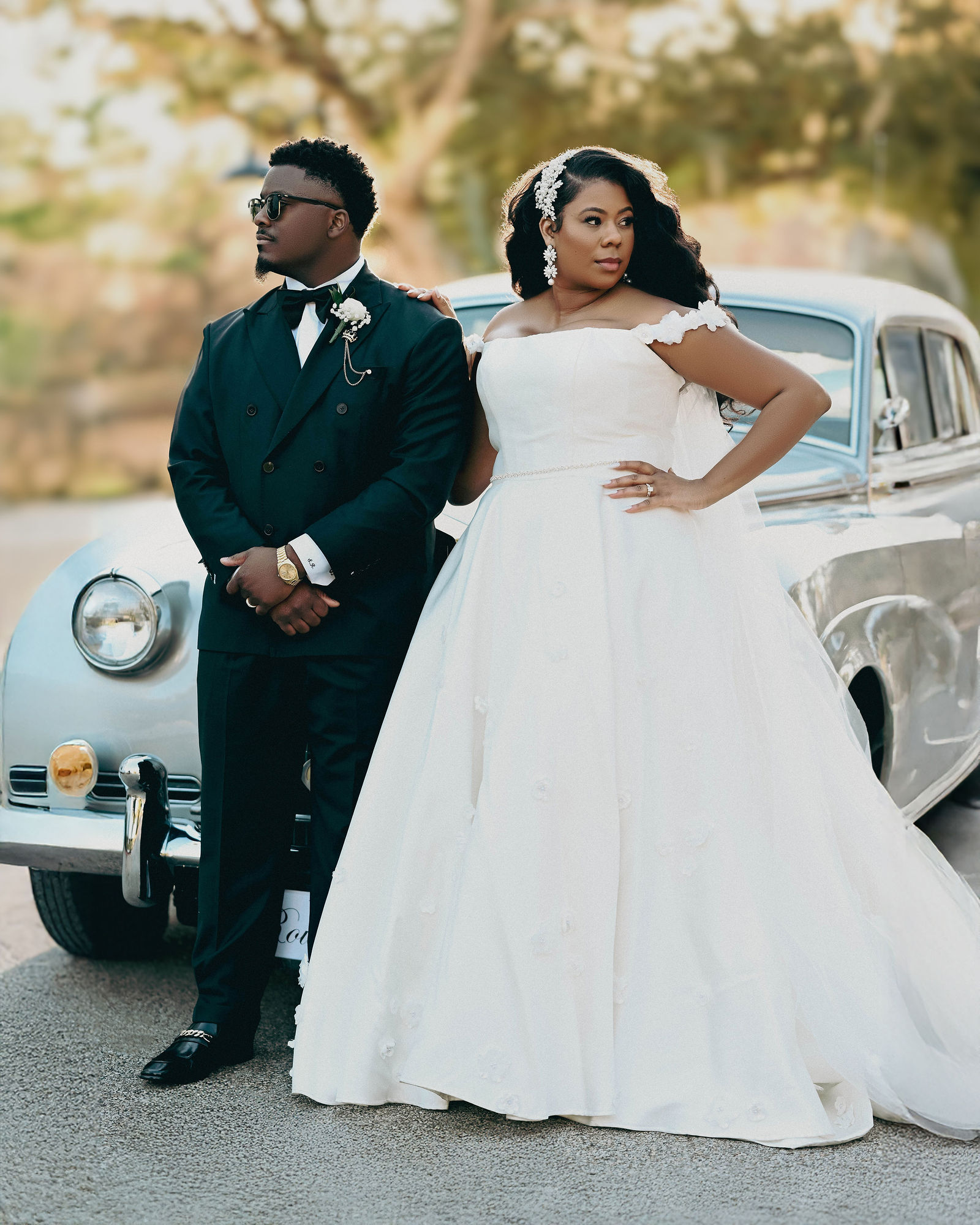 A bride and groom stand outdoors in front of a vintage car, with trees and sunlight in the background.