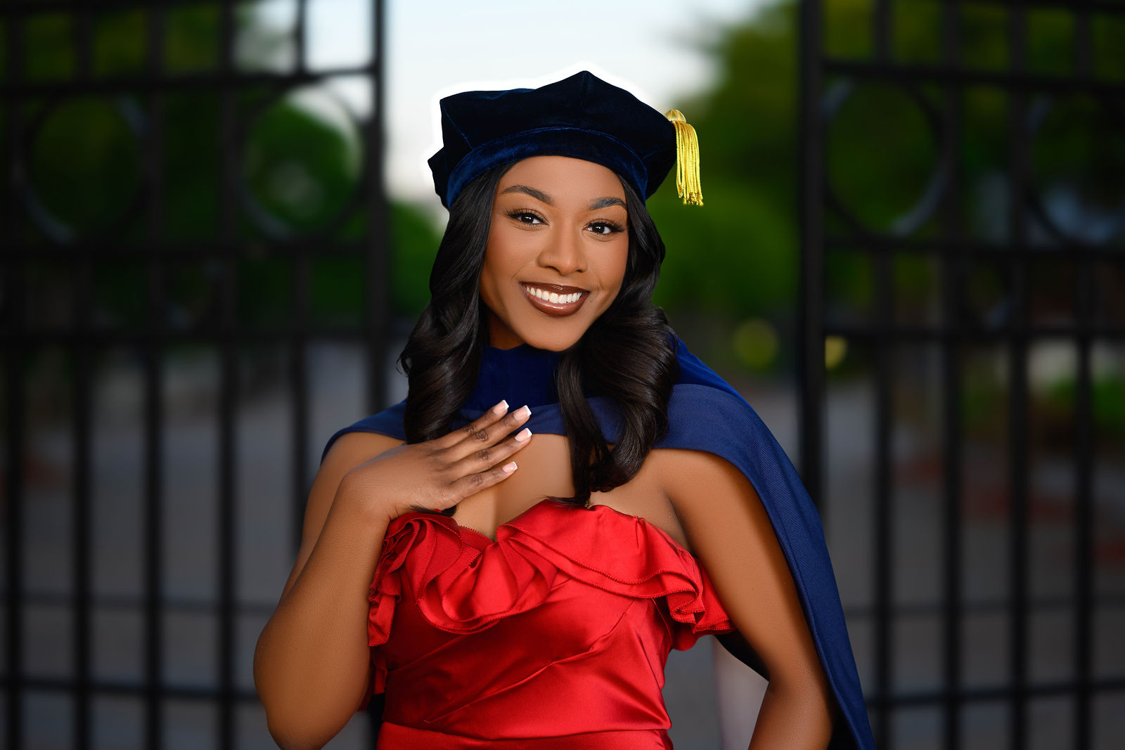 Young woman in graduation cap and gown smiling, holding her hand near her face, outdoors with a gate and greenery behind her.