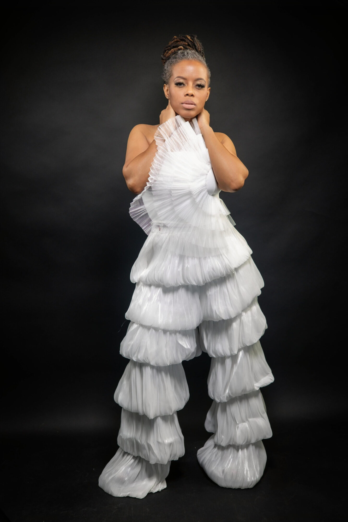 Woman in a white layered dress with ruffles, standing against a black background, looking at the camera with a serious expression.