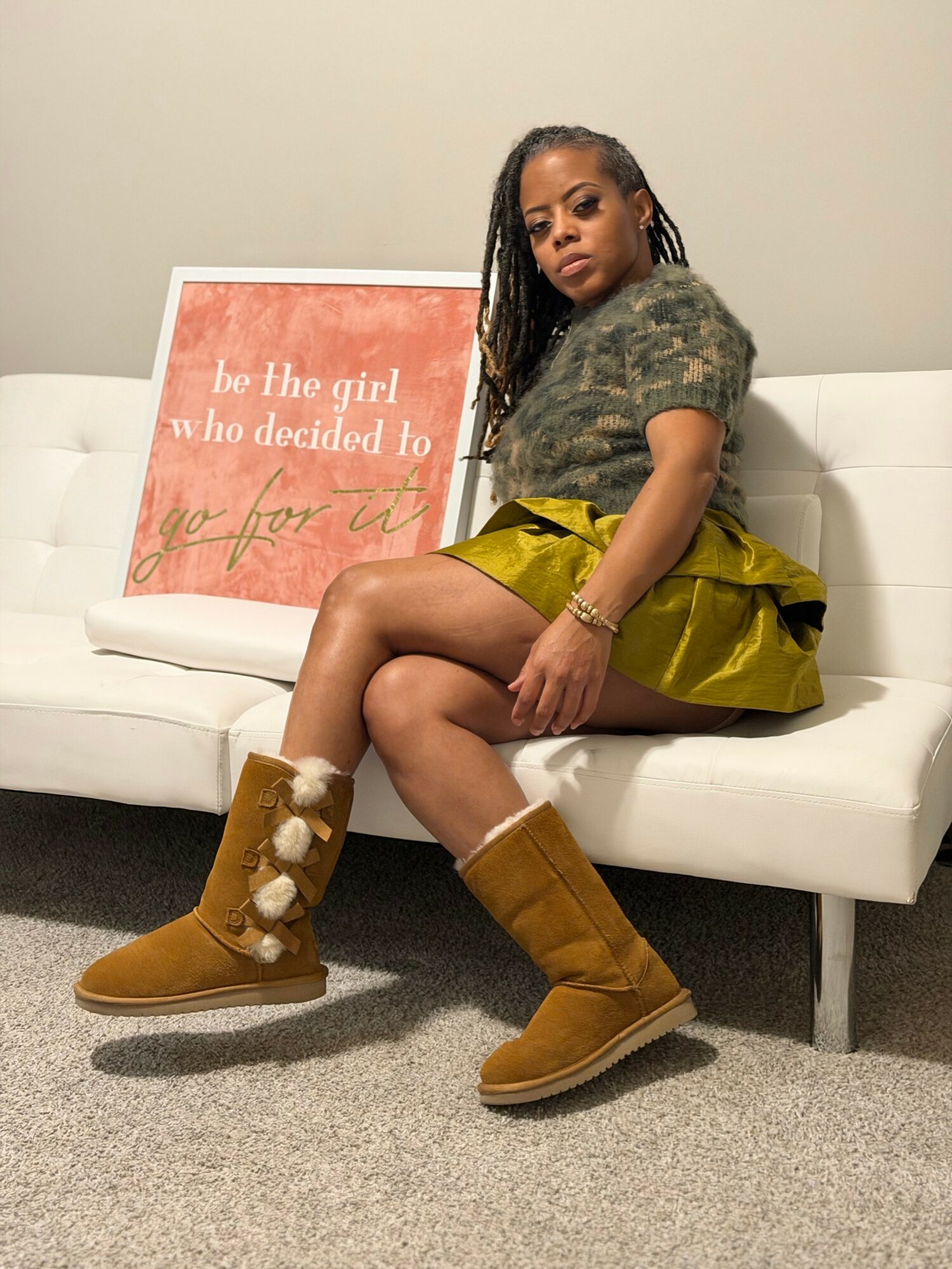 Young woman sitting on a white couch, wearing a camouflage shirt, yellow skirt, and brown boots with pom-poms, next to a pink sign.