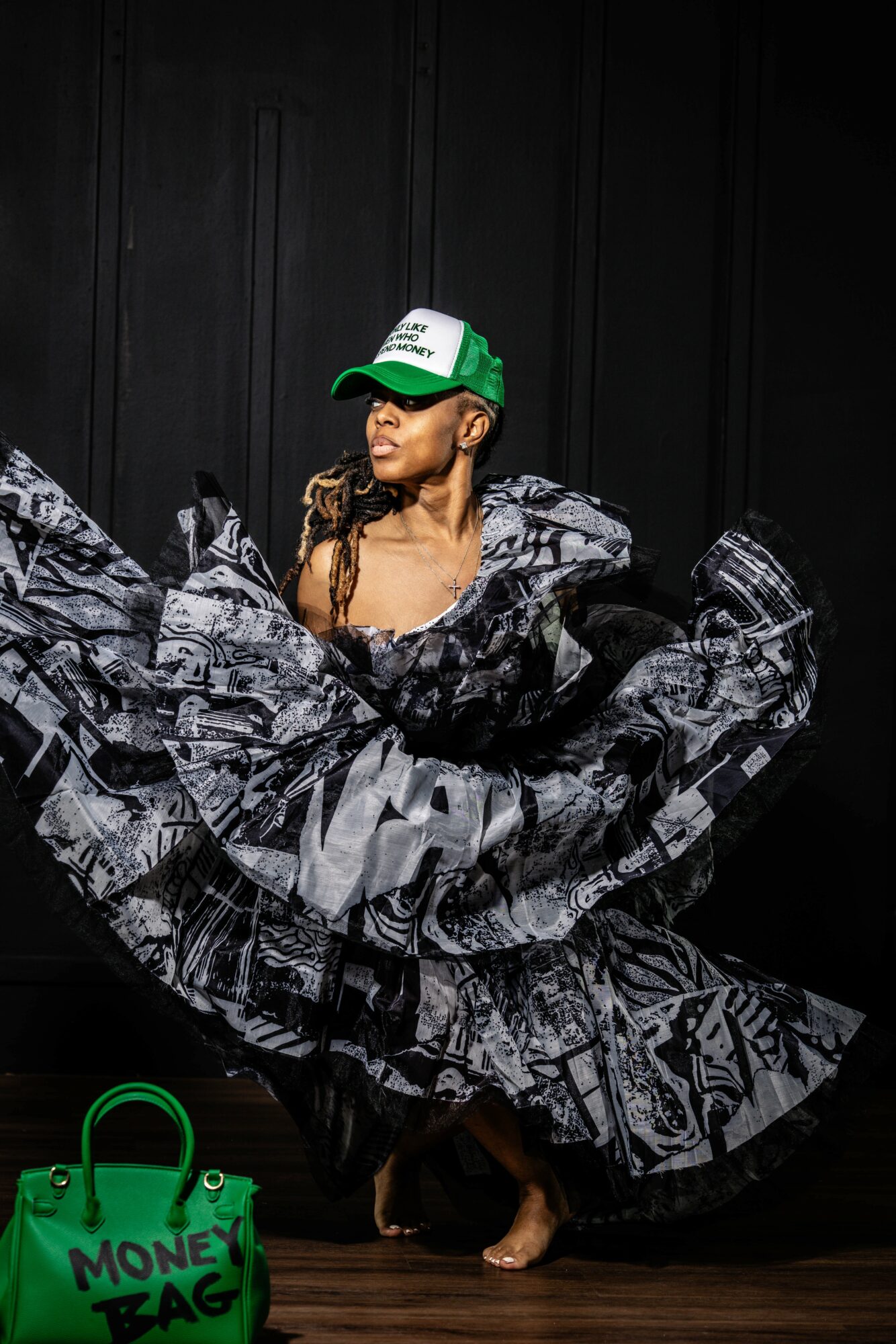 Woman in a black and white patterned dress and green and white cap, standing barefoot against a black background, holding the dress out.