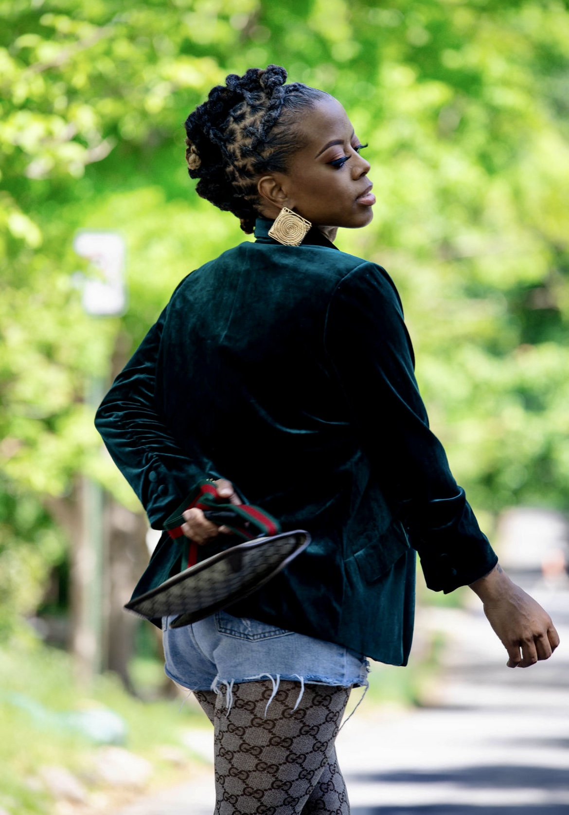 Woman with styled hair and large earrings standing outdoors with green trees in background, holding a small purse.