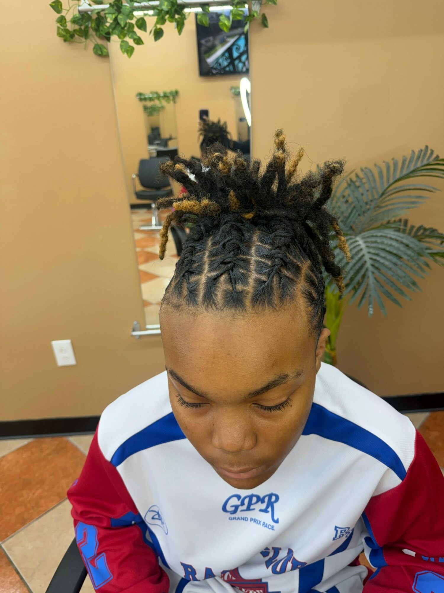 Person with braided hair and dreadlocks, eyes closed, indoors with plants and a mirror in the background.