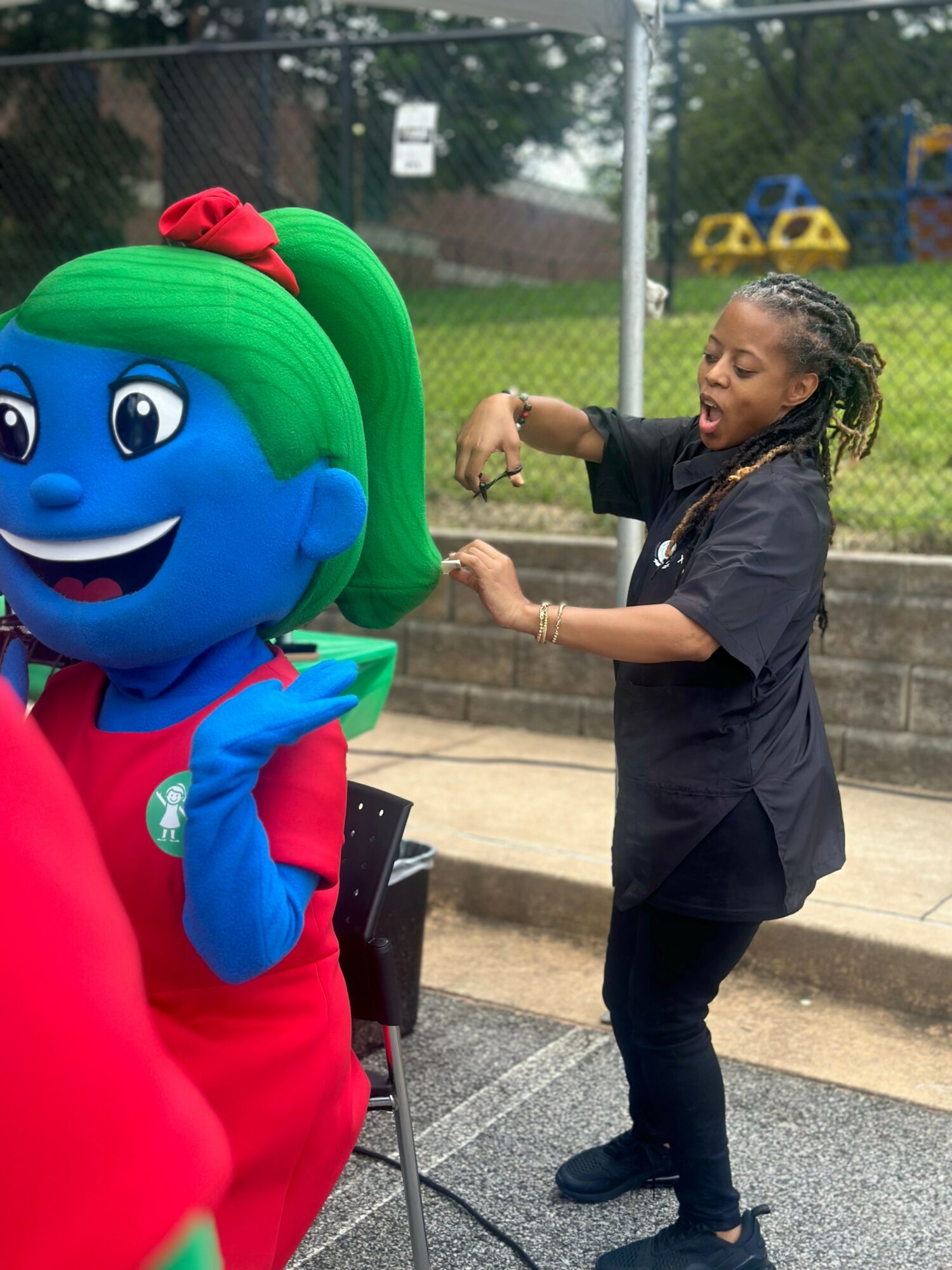 Young girl with braided hair interacts with a large colorful mascot at an outdoor event, with a fence and playground in background.