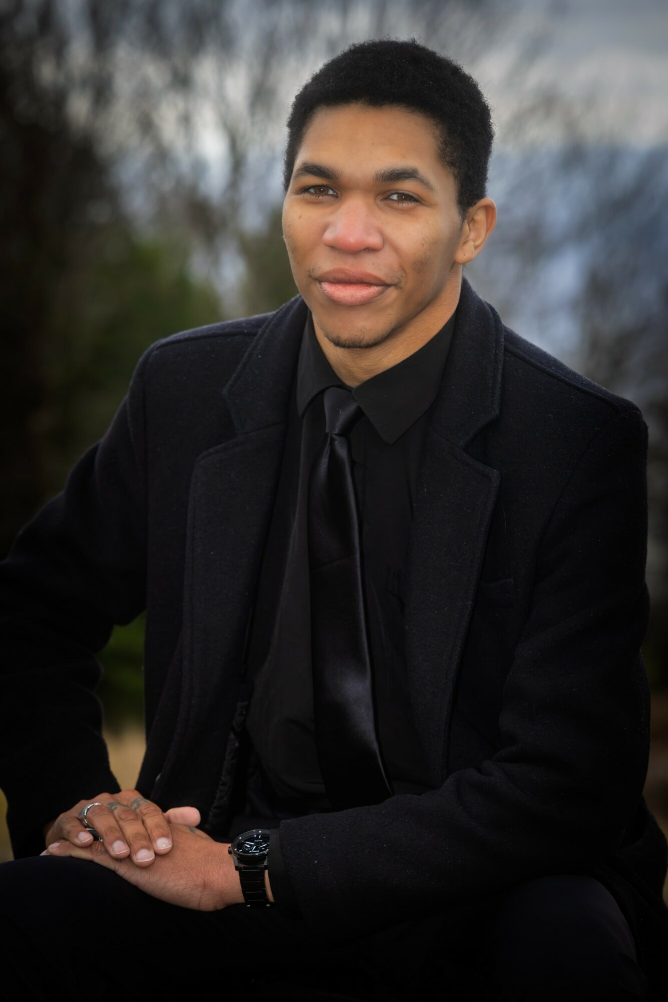Young man in black suit and tie sitting outdoors with trees and sky in background.