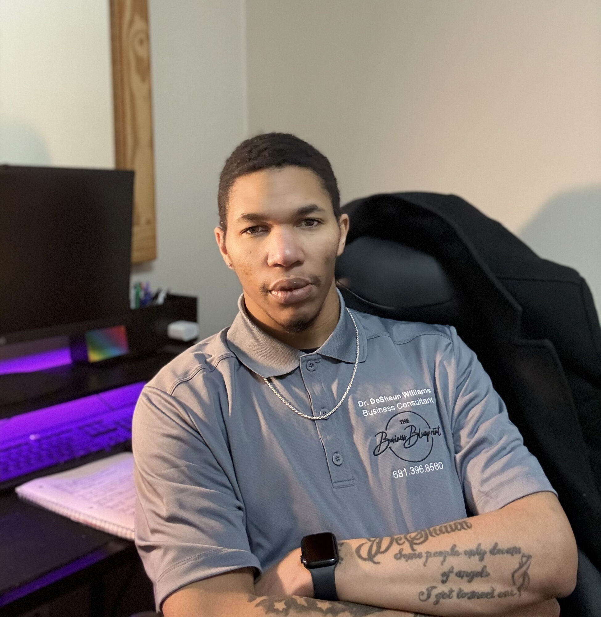 Young man with short dark hair wearing a gray polo shirt, sitting indoors with a computer and notebook behind him.