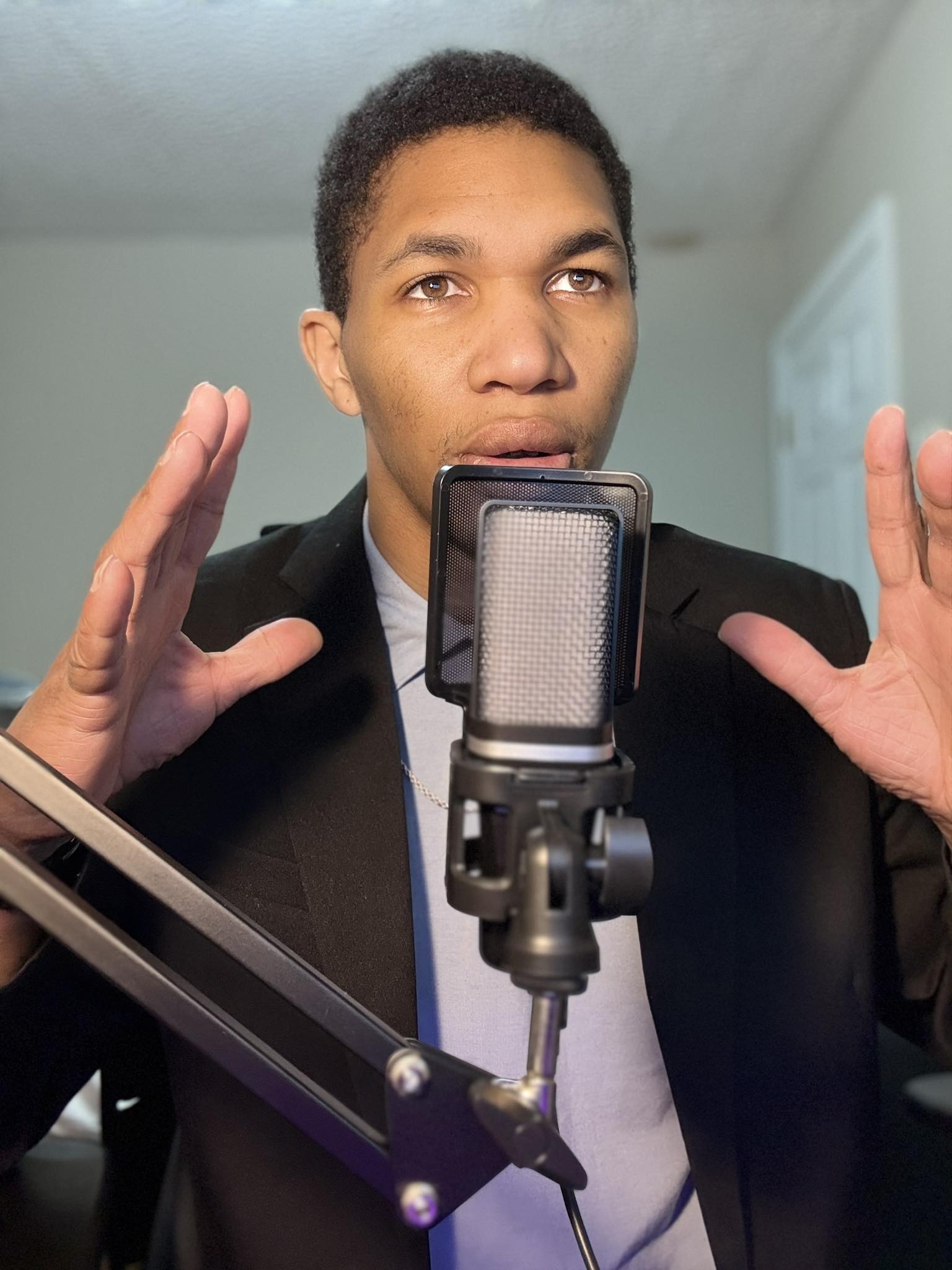 Person with short curly hair in a suit speaking into a microphone, hands raised, in a room with a window.