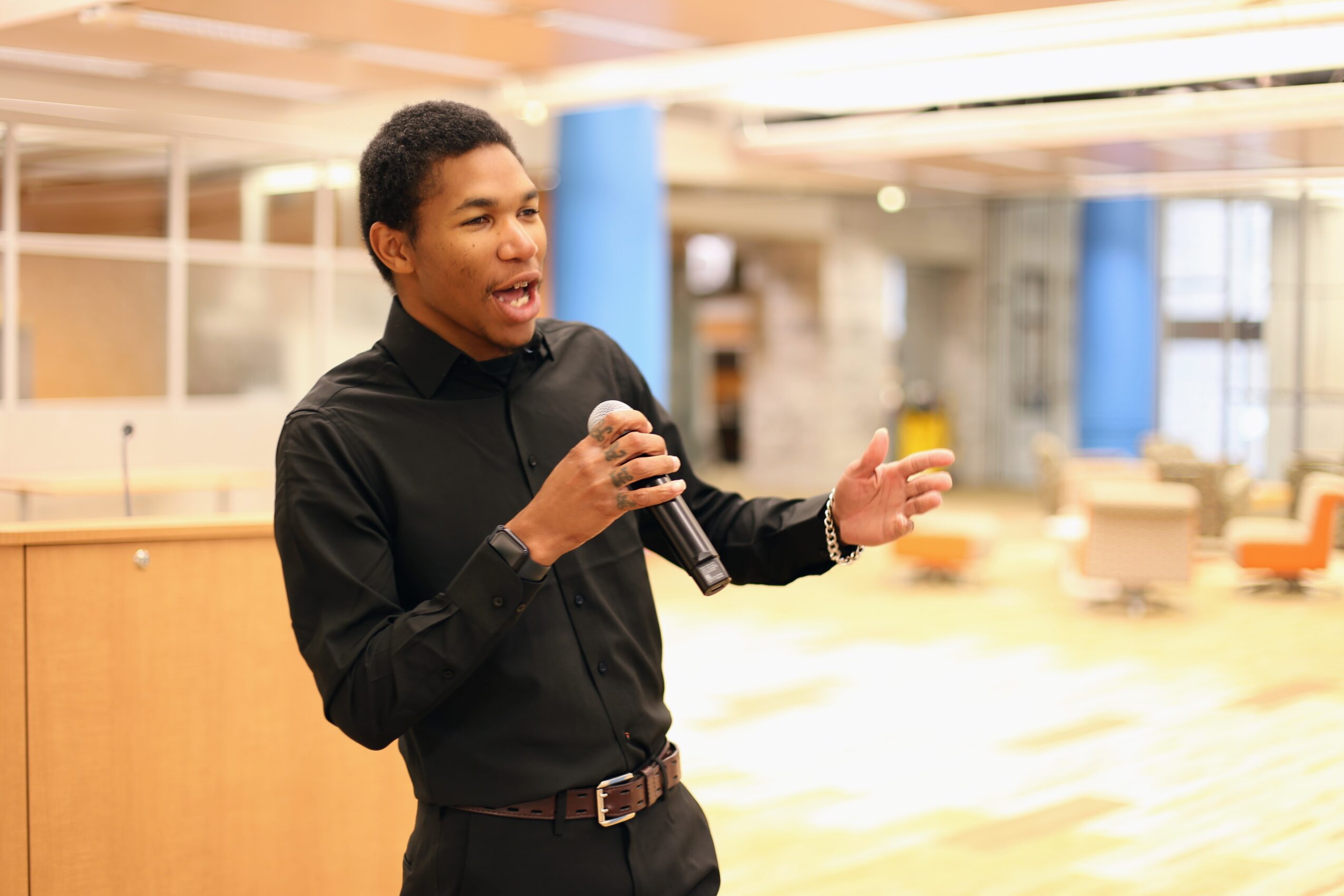 Young man speaking into a microphone in a modern indoor space with seating and large windows.