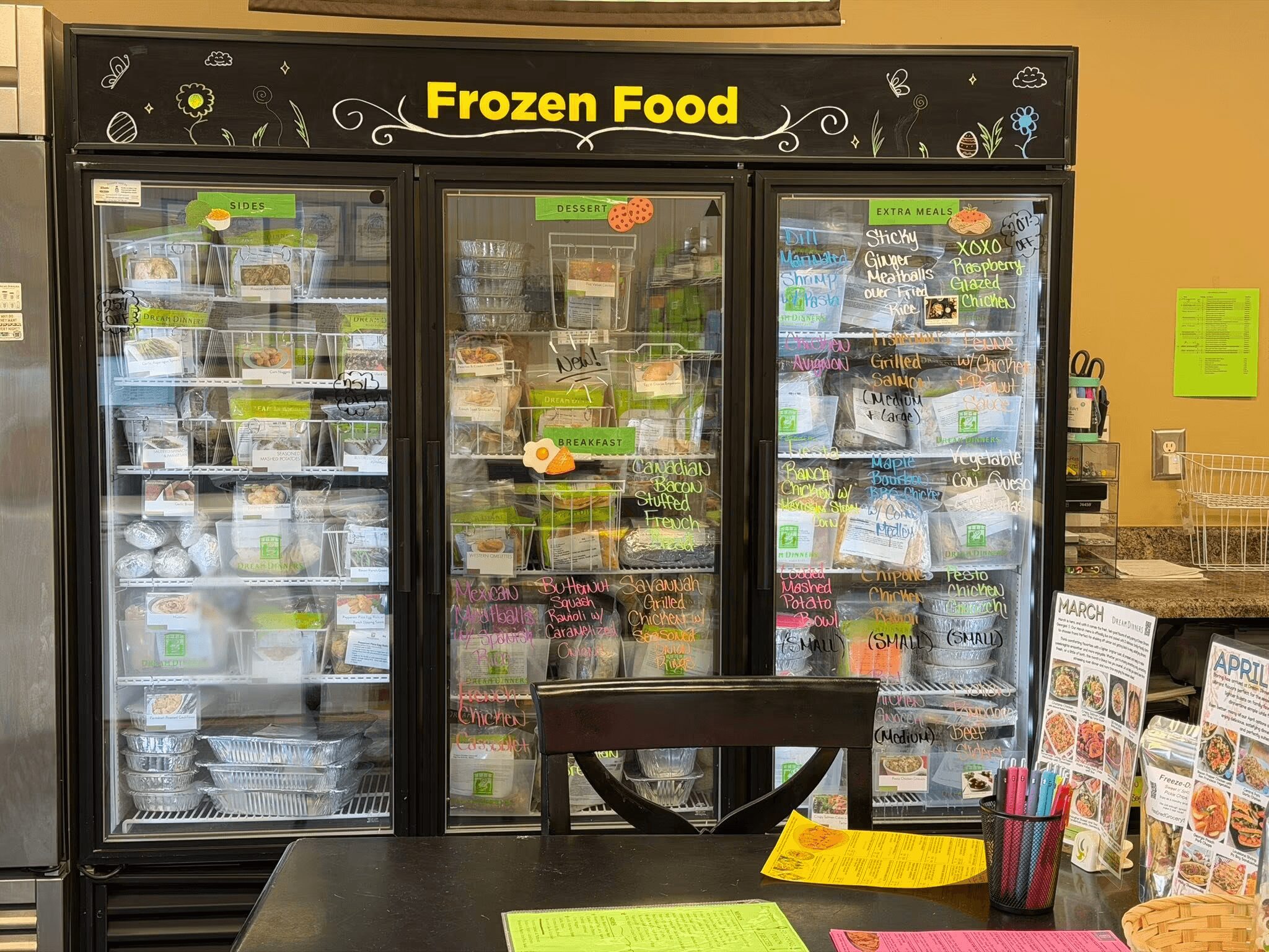 Frozen food freezer with three glass doors filled with various frozen products, black top with yellow 'Frozen Food' sign, in a store.
