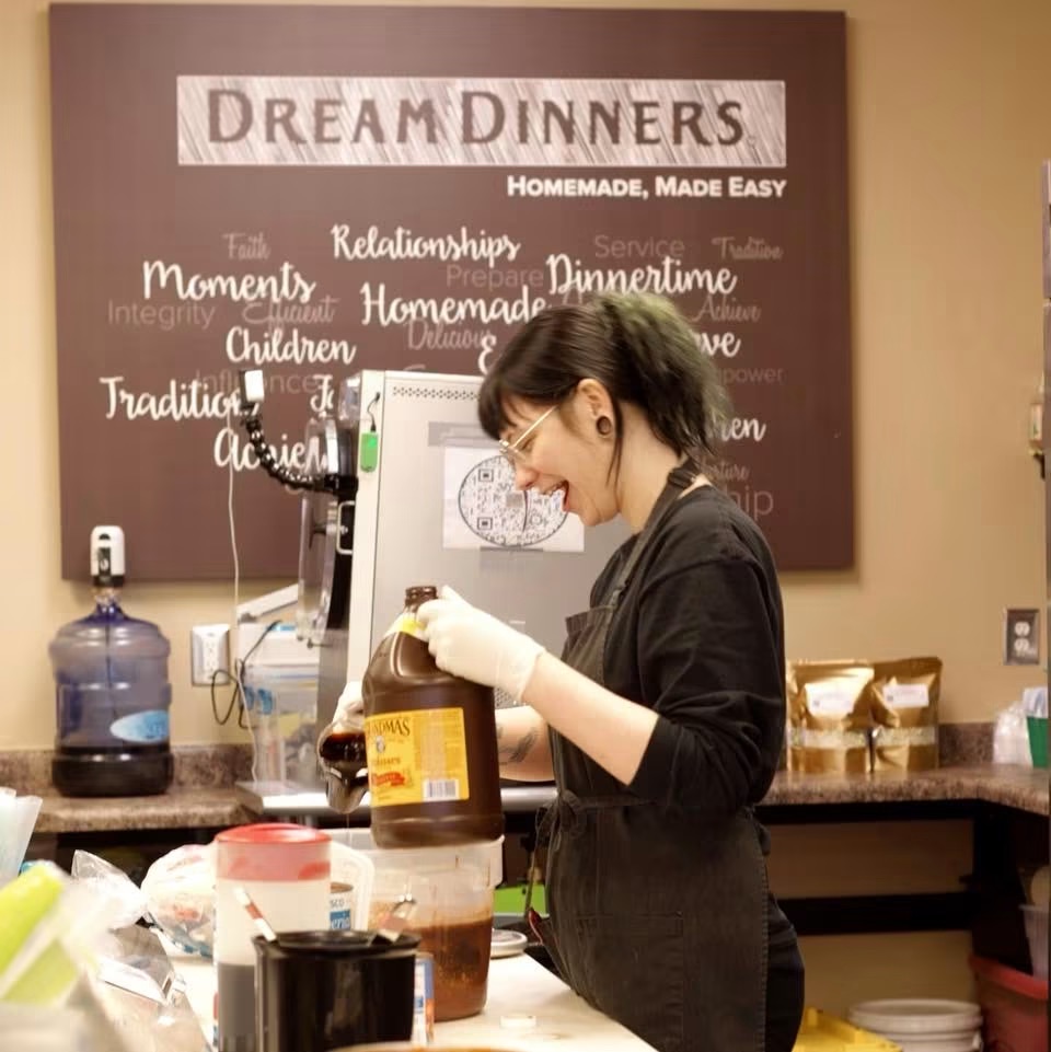Young woman in black apron preparing food in a kitchen, with a chalkboard menu in the background.