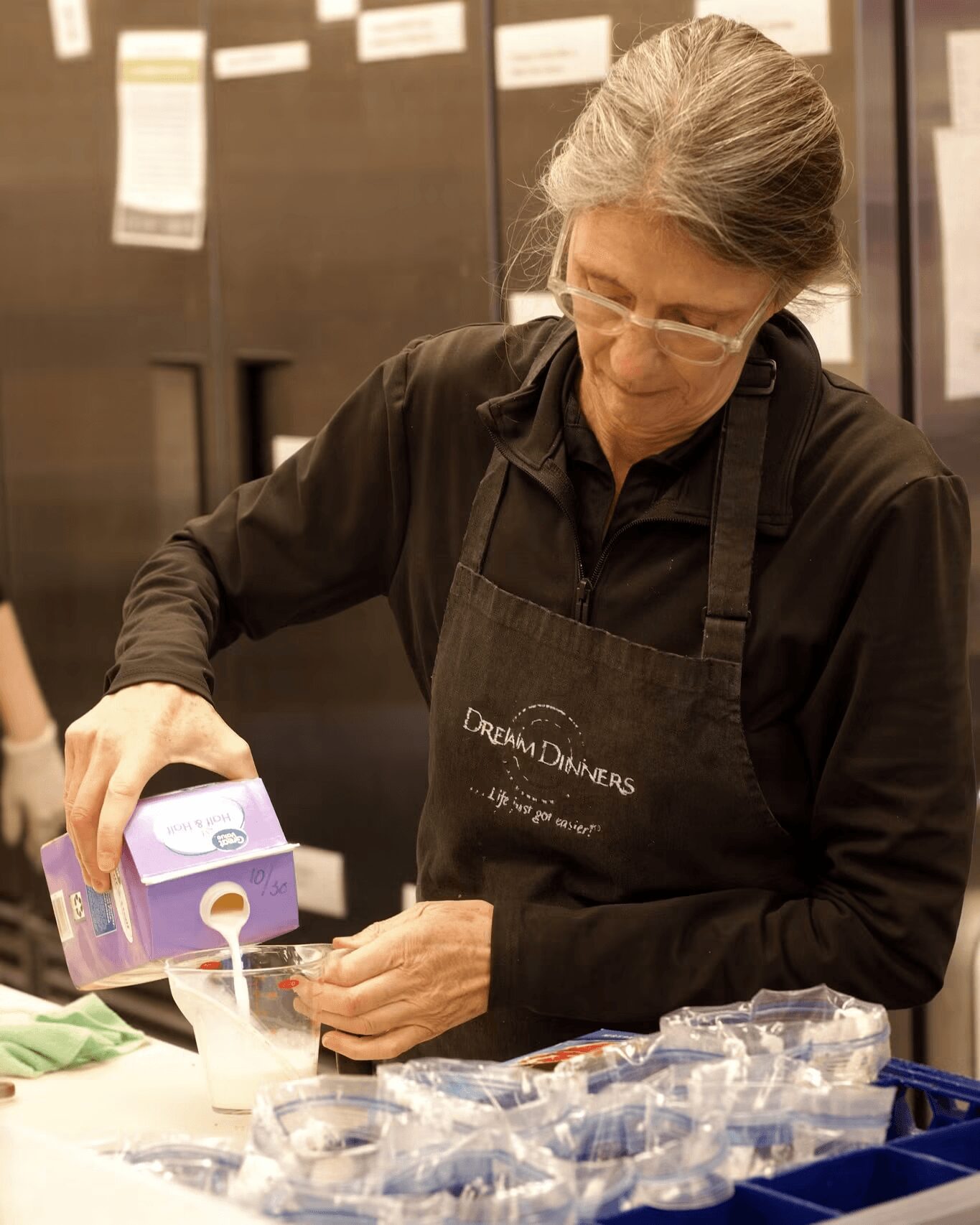 Woman pouring liquid from a purple carton into a small cup, wearing glasses and a black apron, in a kitchen or food prep area.