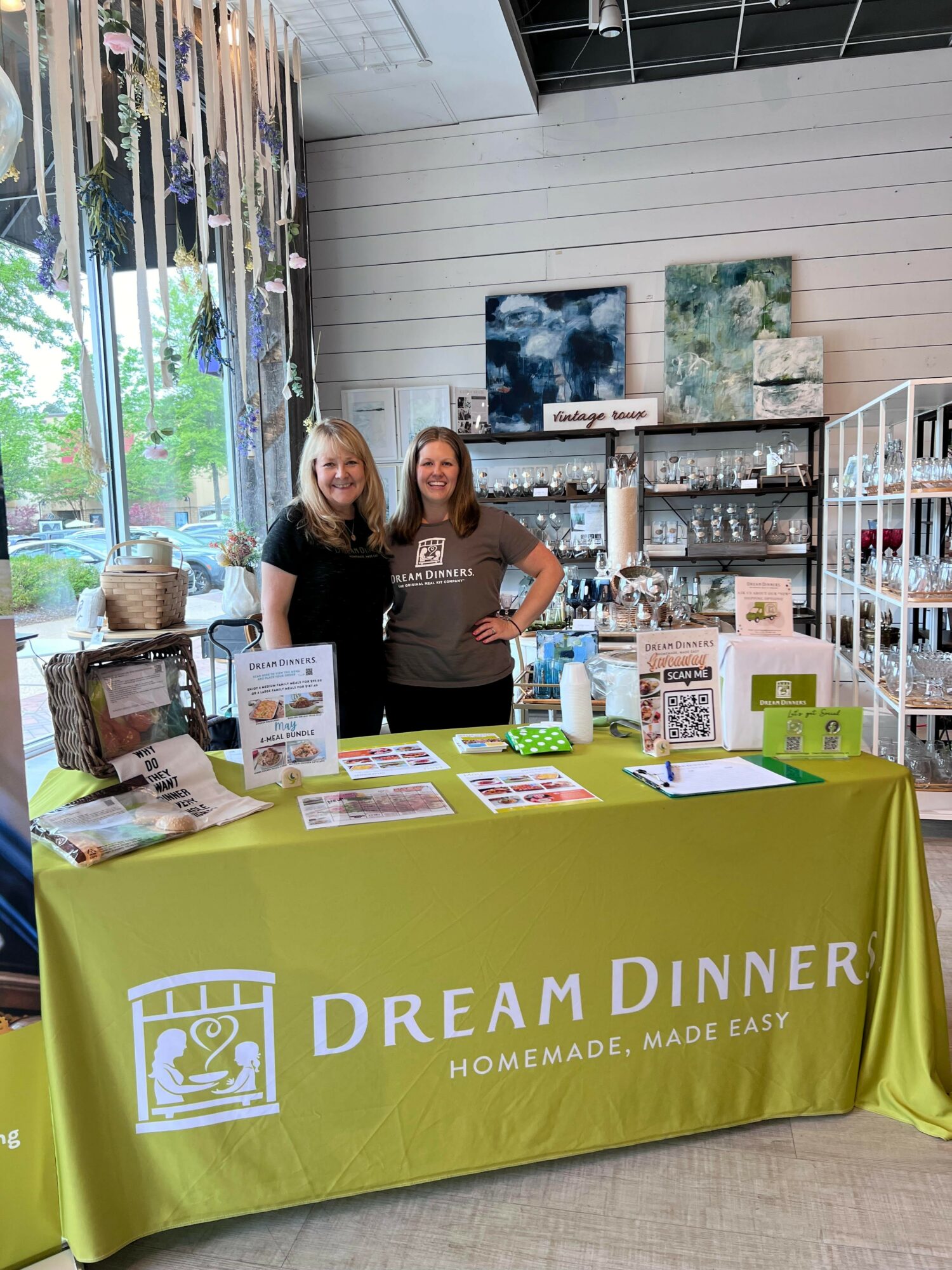 Two women stand behind a table with a green tablecloth displaying 'Dream Dinner' logo, in a store with shelves and artwork.