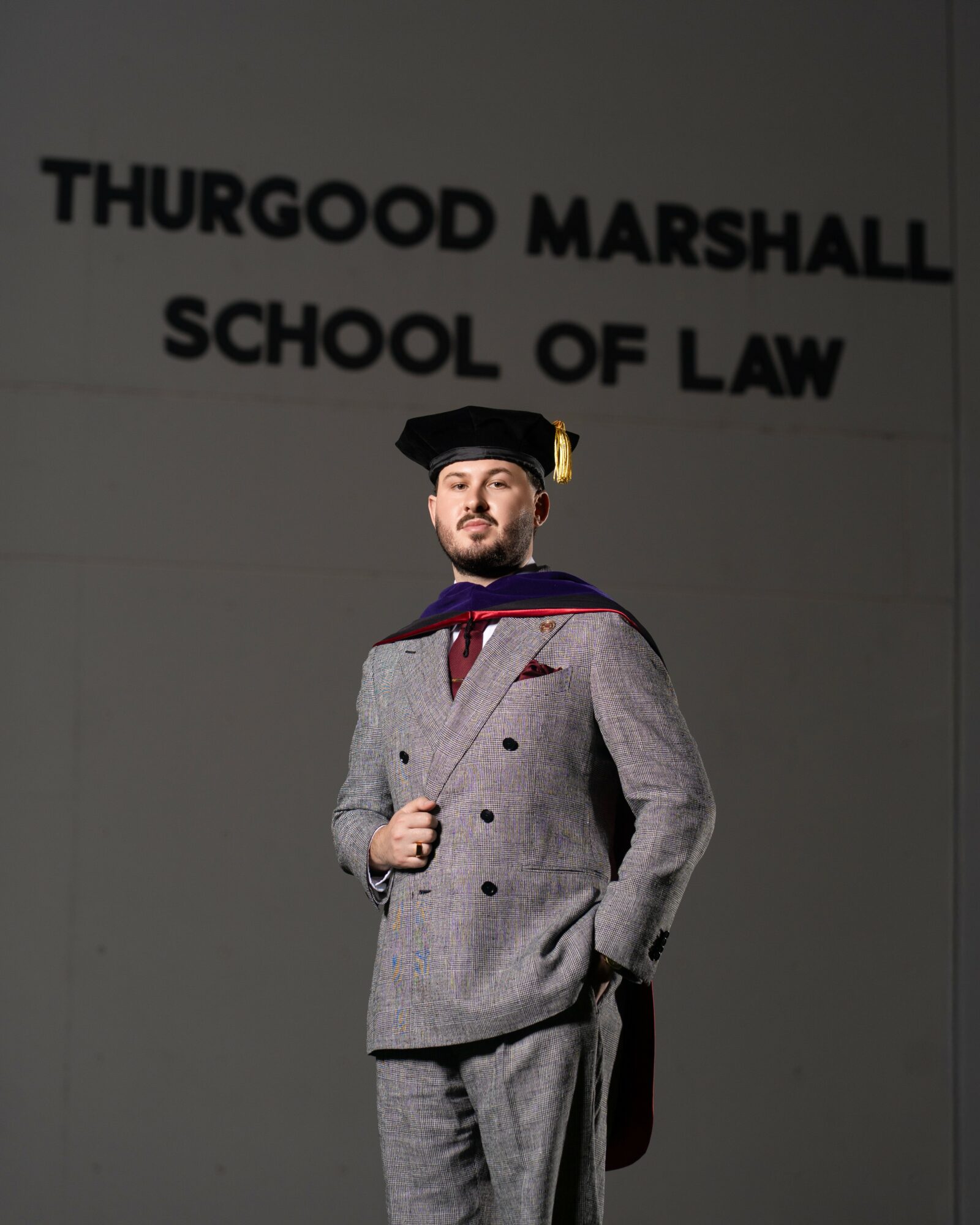 Graduate in a gray suit and cap standing in front of a wall with text, holding a diploma. The wall reads 'THURGOOD MARSHALL SCHOOL OF LAW'.