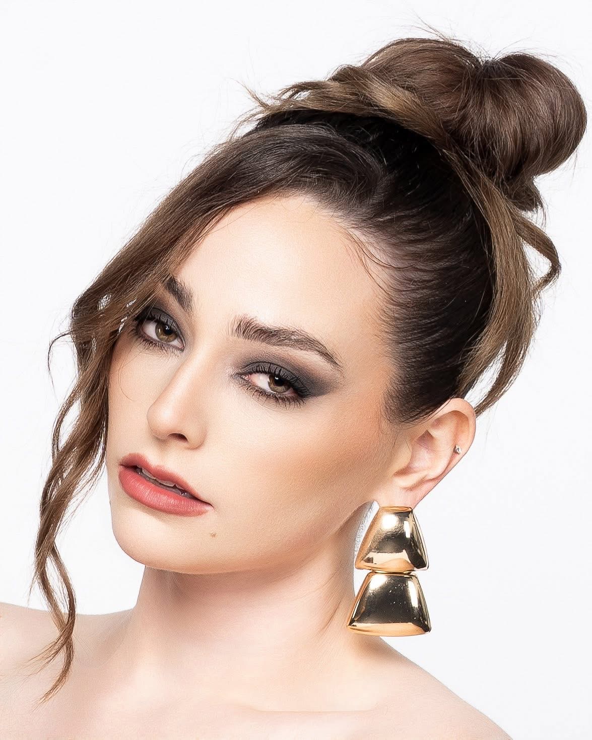 Woman with styled hair, makeup, and large earrings, looking at the camera against a white background.