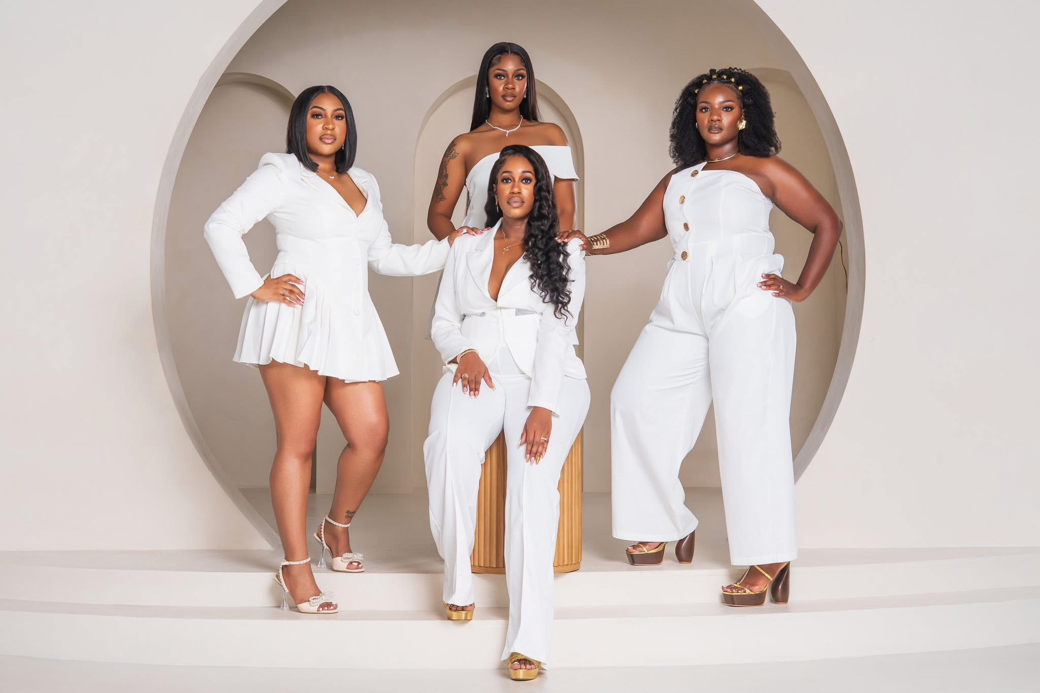 Four women in white outfits pose inside a circular opening, with two standing and two sitting on a chair.