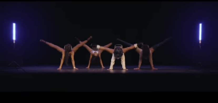 Five dancers perform a pose on stage with dark background and vertical blue lights, arms and legs extended outward.