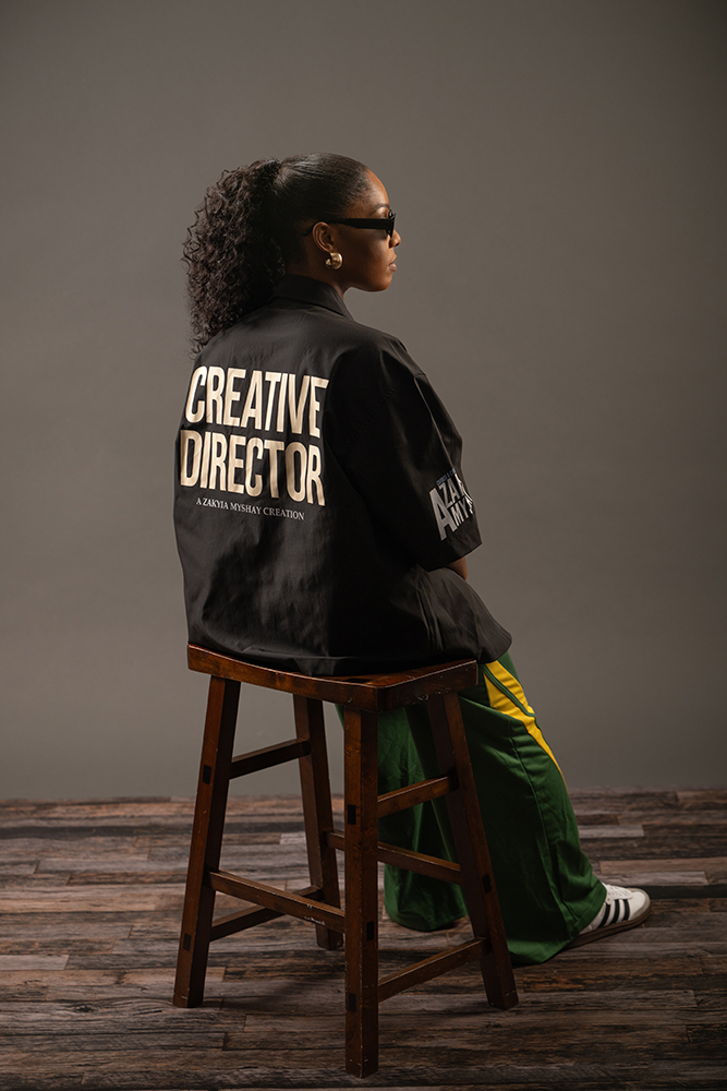 Woman with curly hair and sunglasses sitting on a wooden stool, wearing a black jacket with white text, green pants, and sneakers.