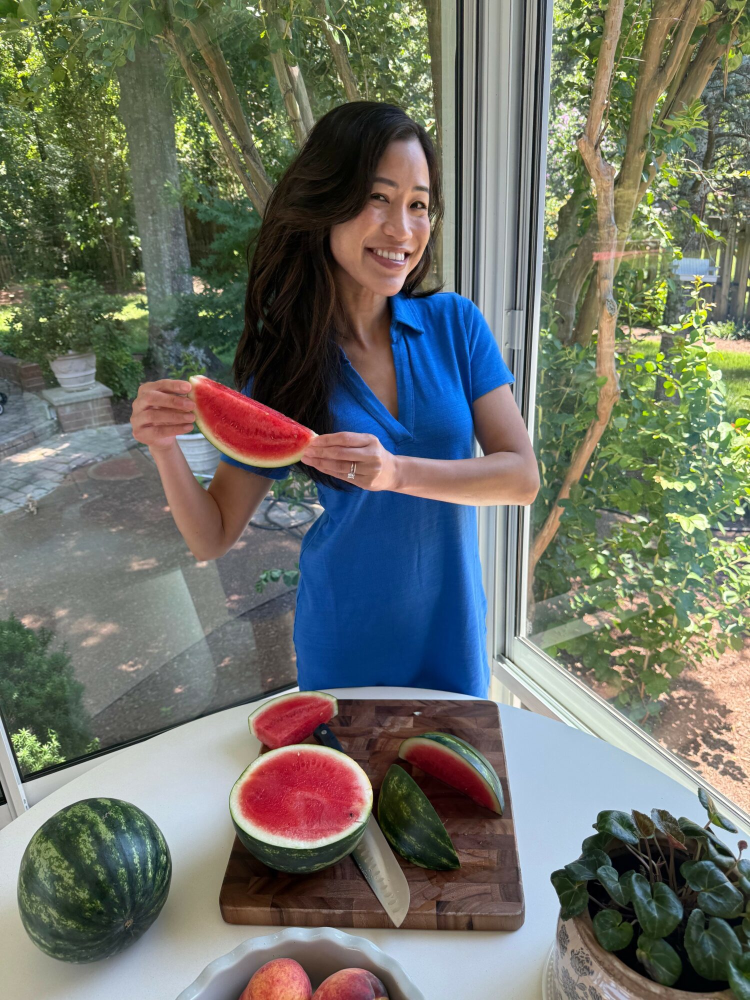 Woman in blue dress holding a watermelon slice near a window with trees outside.