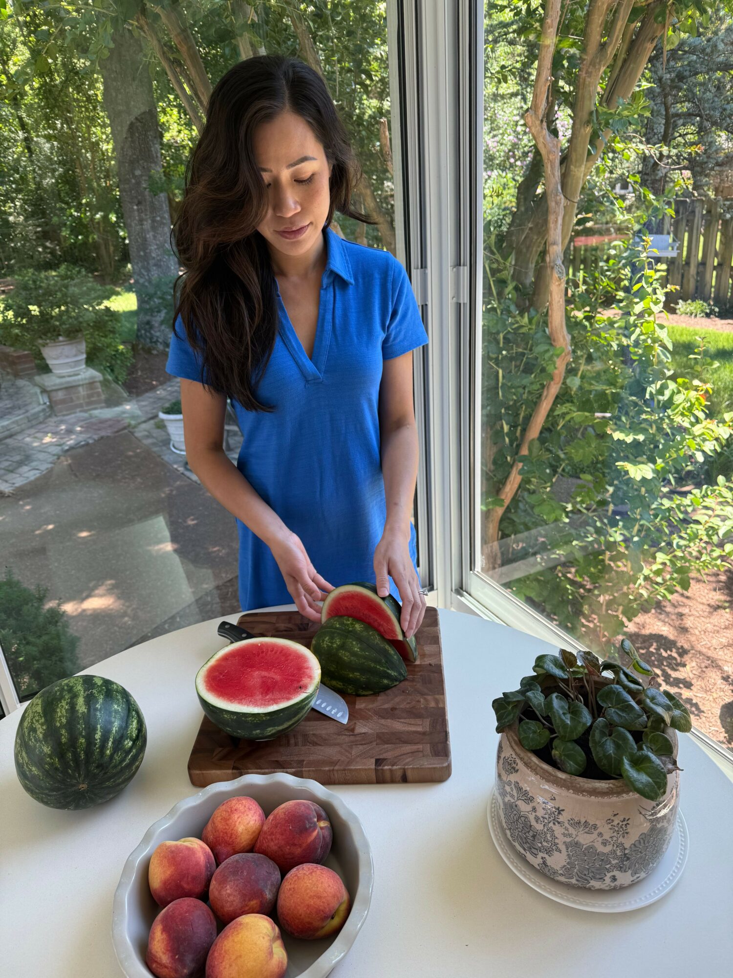 Woman in blue dress standing next to a table with watermelons, peaches, and a potted plant, outdoors near a window.