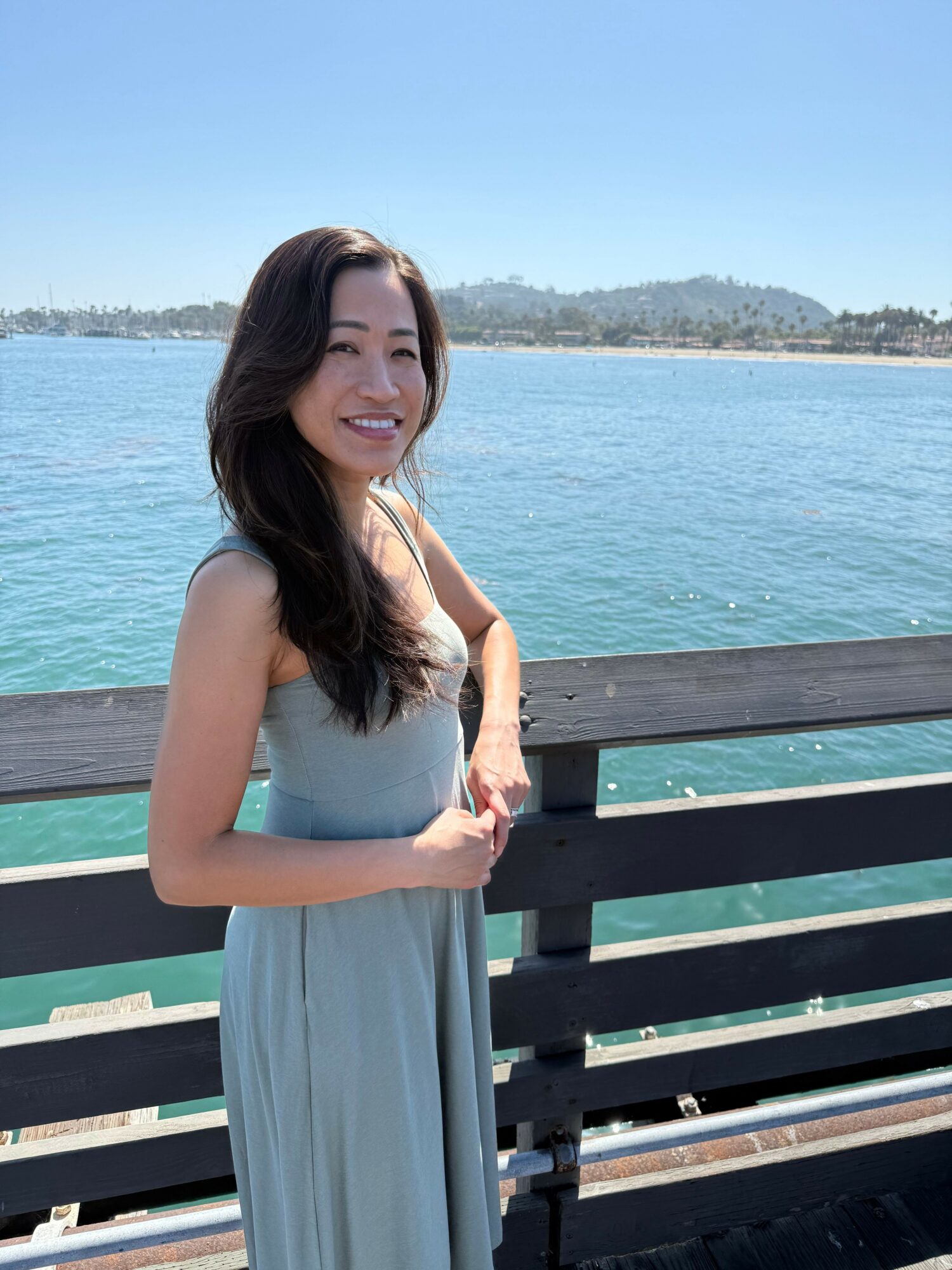 Woman in a light blue dress standing on a pier with water and land in the background, smiling, sunny day.