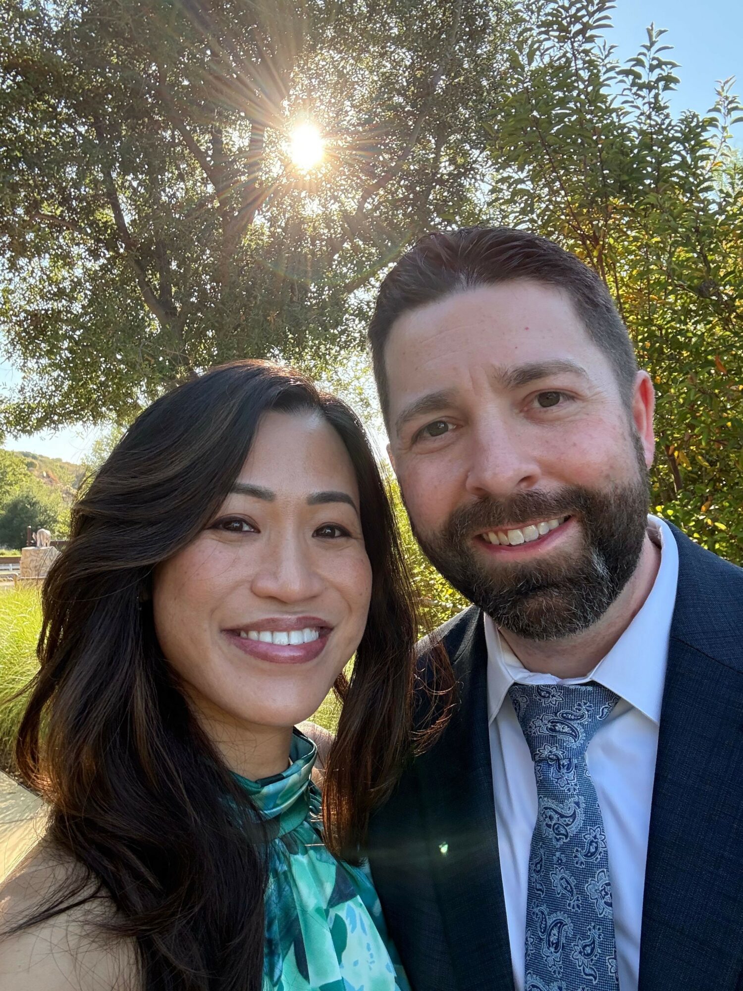 Two people smiling outdoors with sunlight and trees in the background.