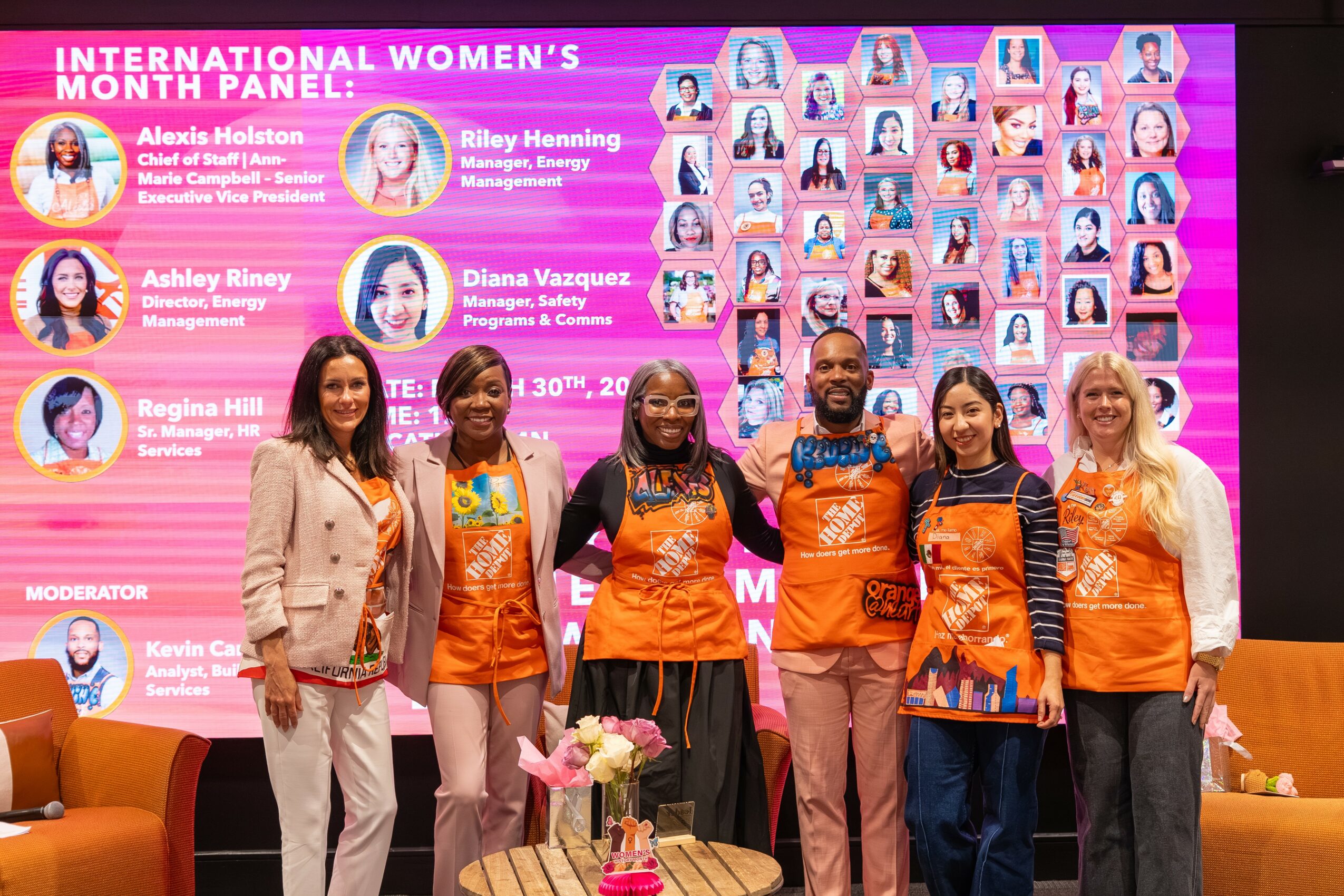 Six women standing on stage in front of a pink backdrop with photos and text, some wearing orange shirts, holding flowers.