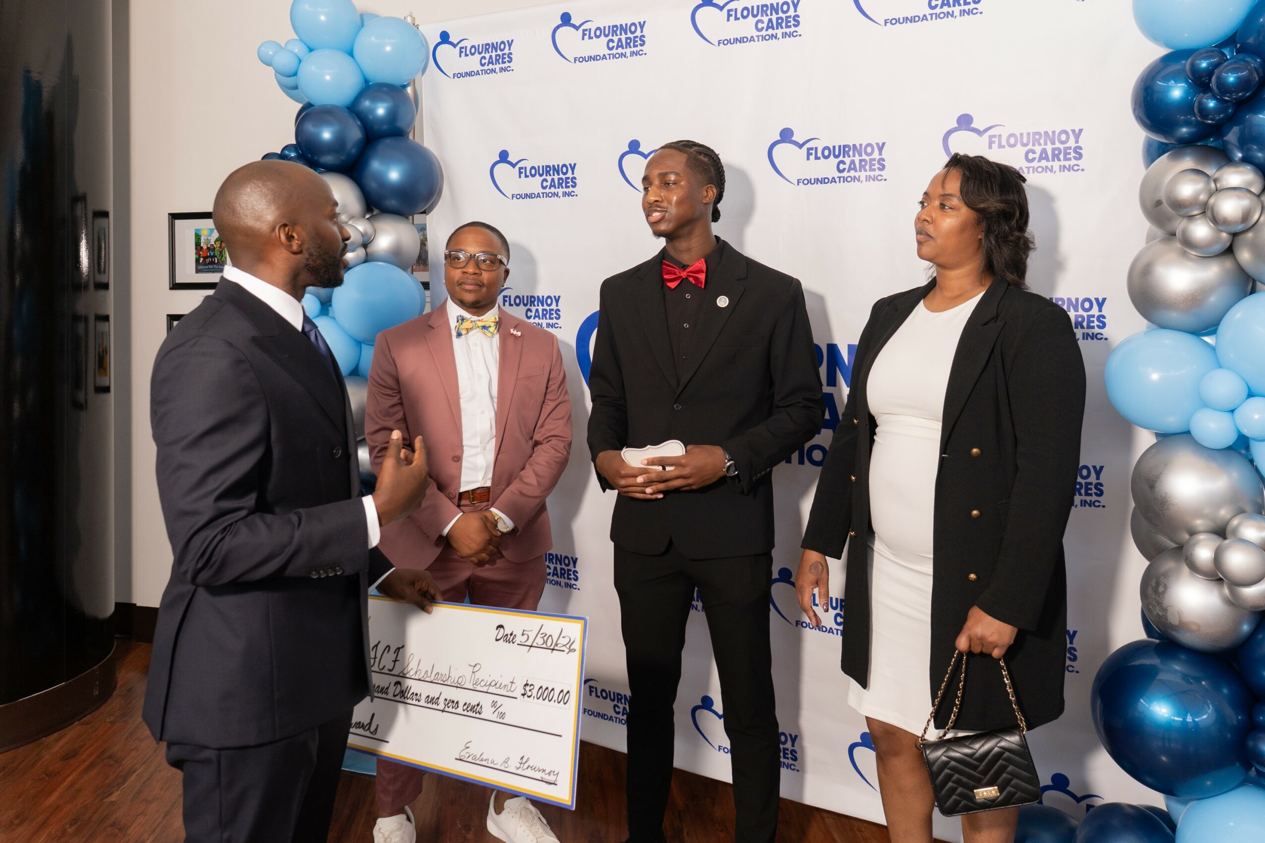 Four people in formal attire standing and talking at an event with blue, silver, and black balloon decorations and a branded backdrop.