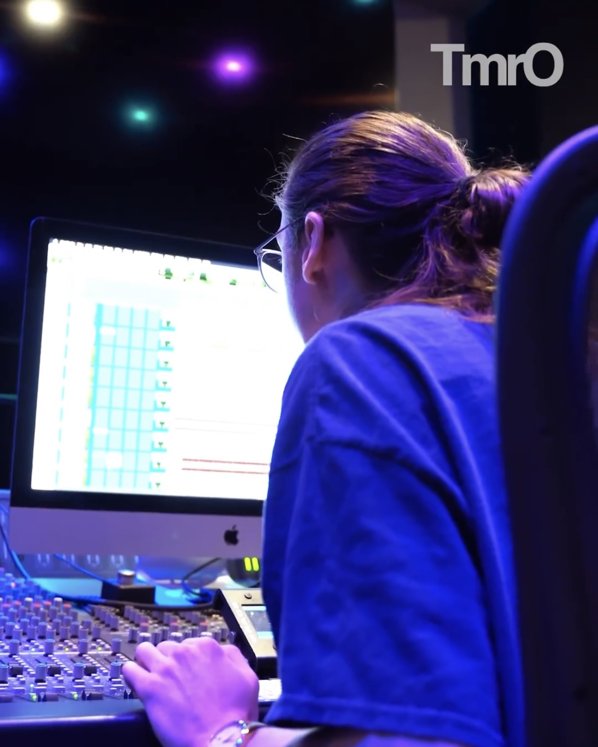 Person with hair in a ponytail working on a computer with a large screen, in a dark room with colorful lights
