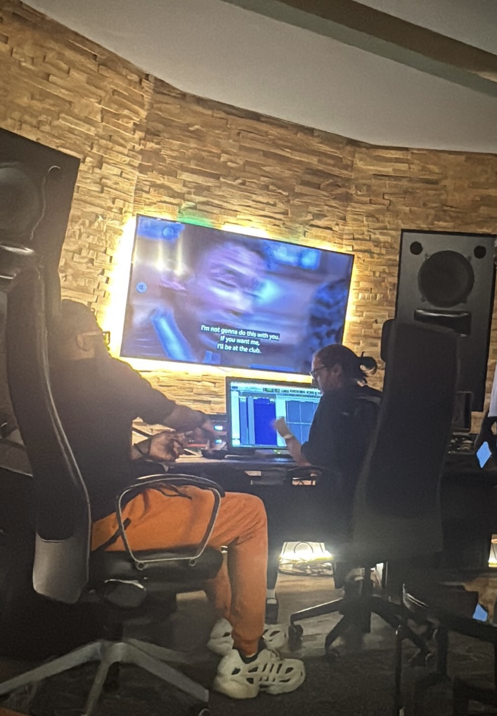 Two people working at a music production studio with a large screen displaying a face and subtitles, surrounded by sound equipment.