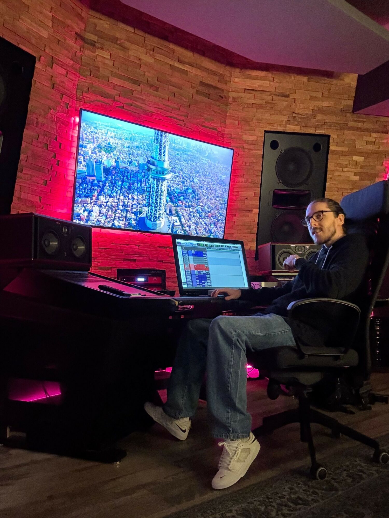 Person sitting at a desk with a large screen, laptop, and speakers in a room with wooden walls and pink lighting.
