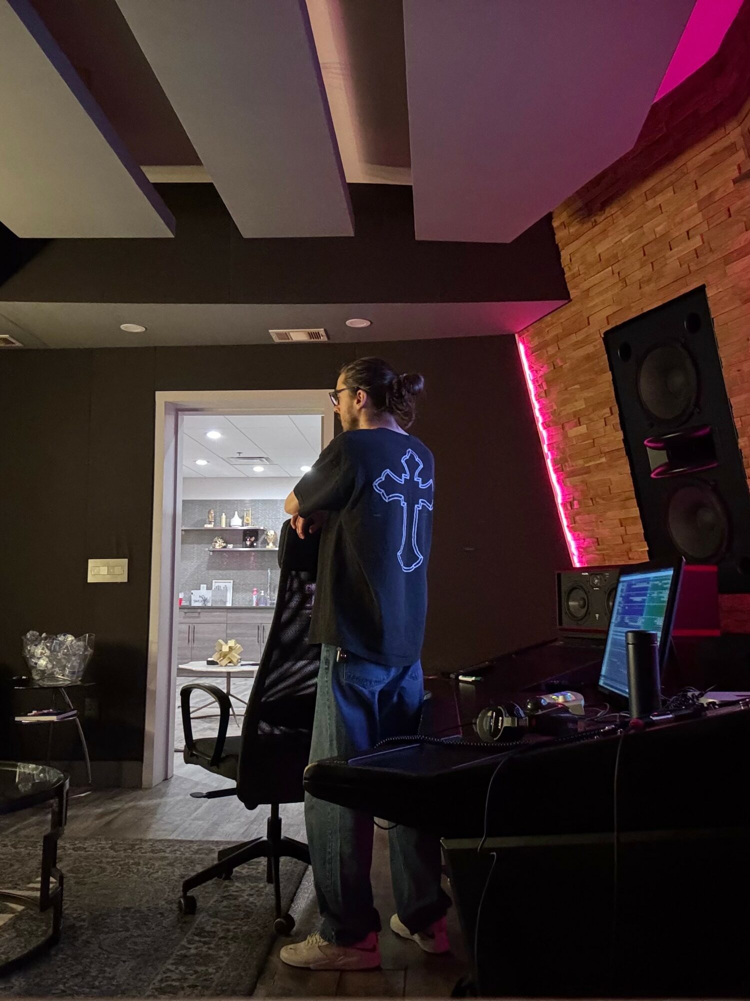 Person with long hair and glasses stands in a room with black walls, a brick wall, and a computer setup, facing a mirror.