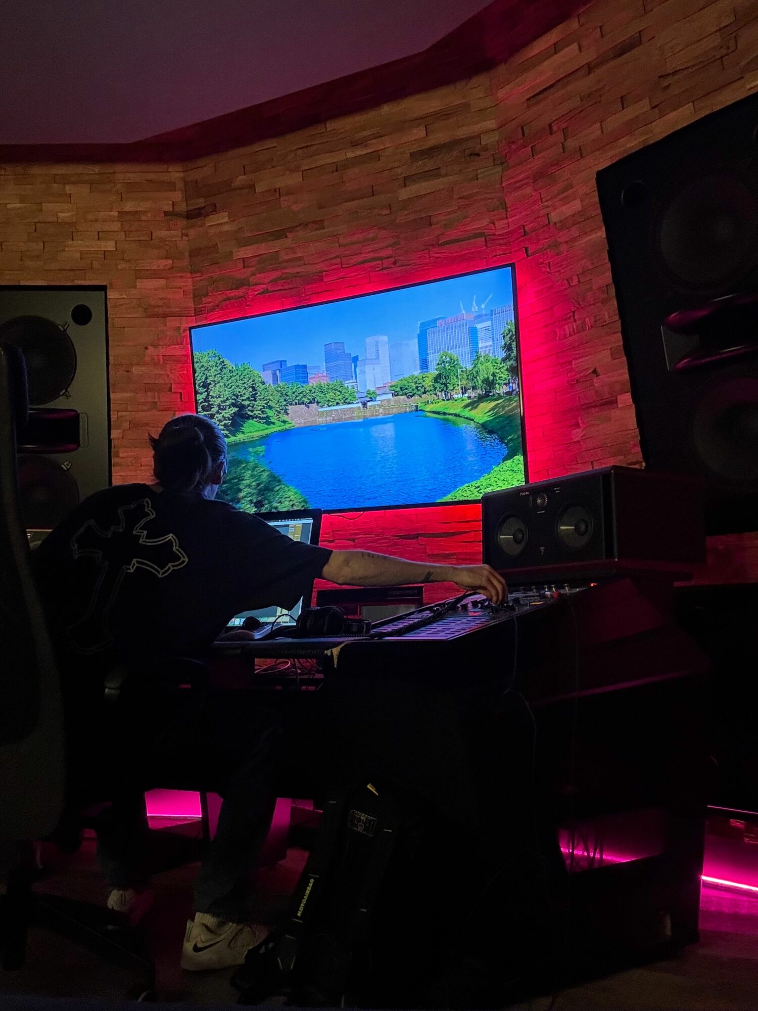 Person working at a computer in a room with a large screen displaying a landscape scene, surrounded by speakers and equipment.