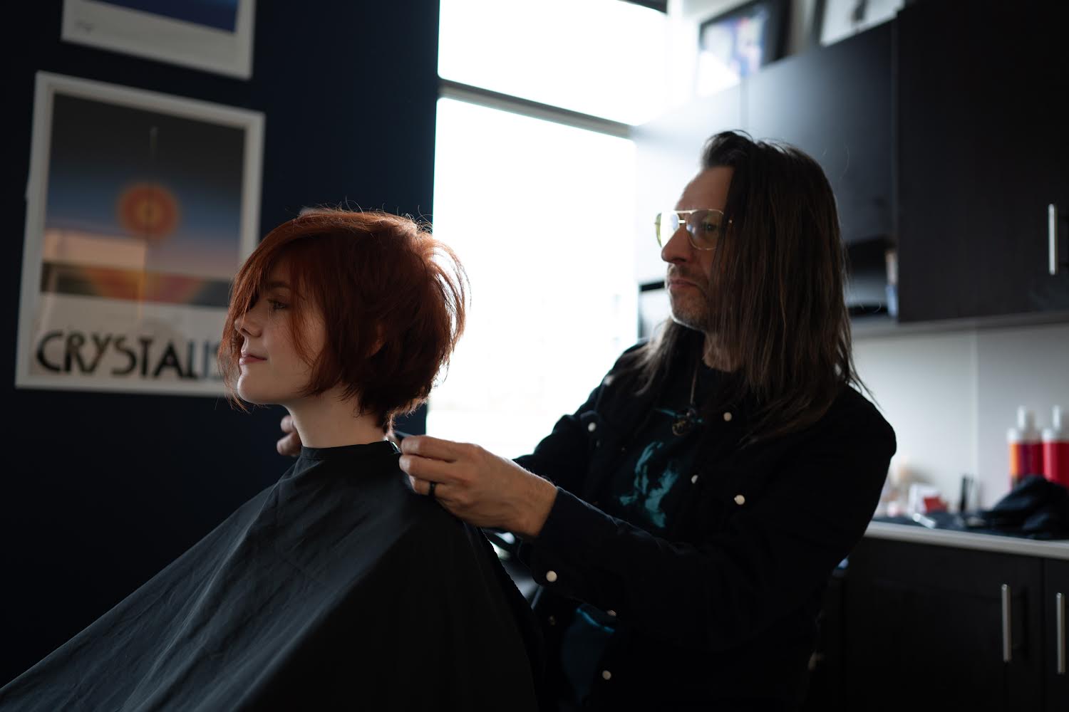 A hairstylist works on a woman's hair in a salon with dark walls and bright window light.