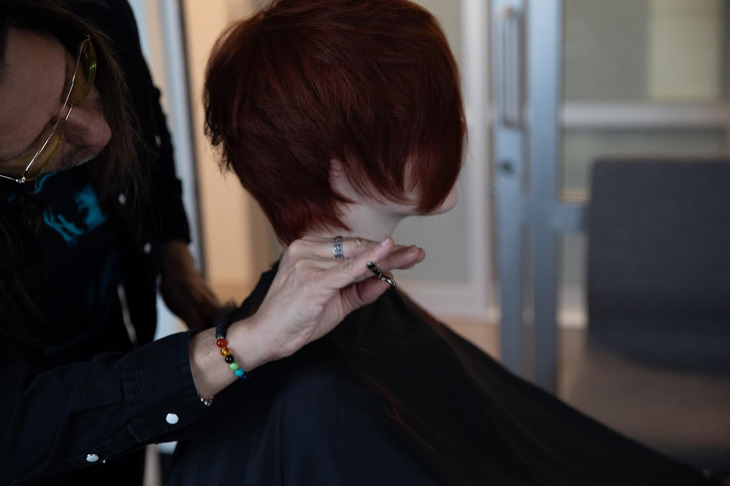 Person with short red hair getting a haircut in a salon, with a stylist working on their hair.