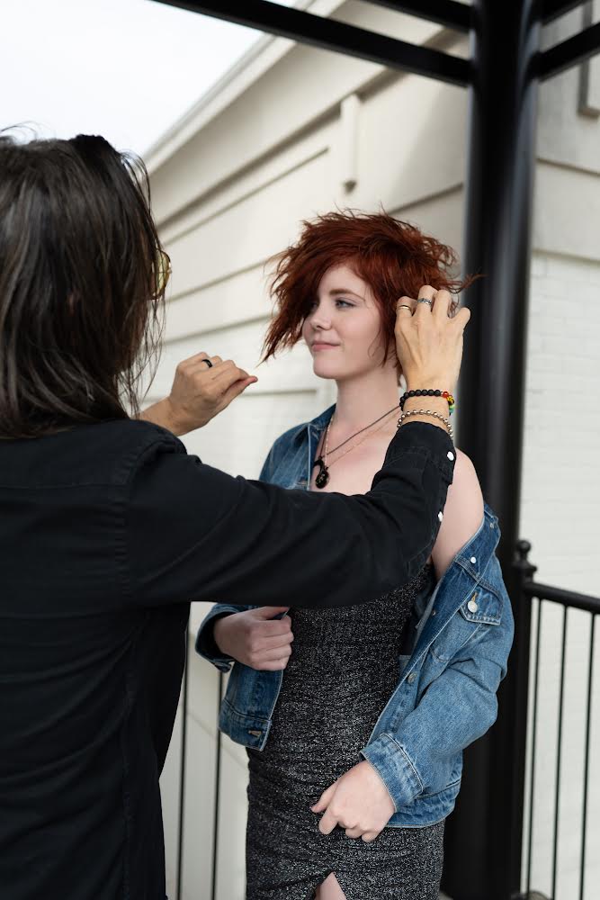 Two women are preparing for a photo, one adjusting the other's hair outdoors near a white building and black railing.