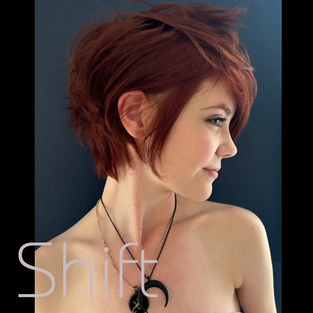 Side profile of a woman with short, reddish-brown hair, wearing a necklace with a crescent moon pendant, against a dark background.