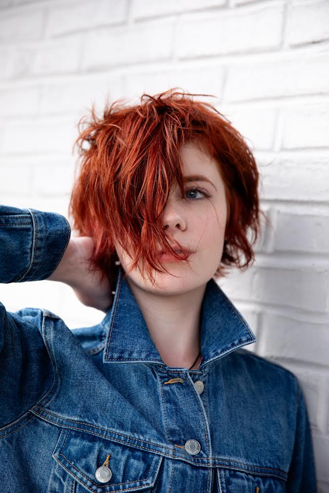 Young woman with short, curly red hair wearing a denim jacket, standing against a white brick wall.