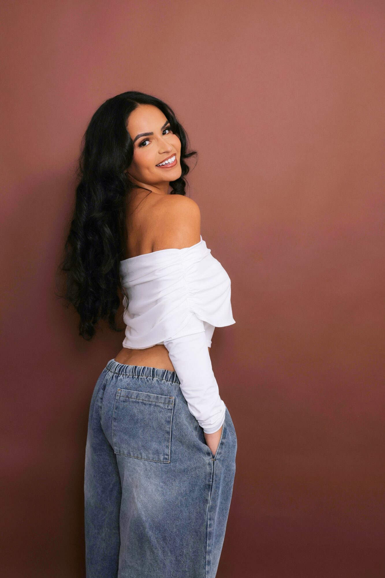 Young woman with long dark hair smiling, wearing an off-shoulder white top and high-waisted jeans, standing against a pinkish-brown background.