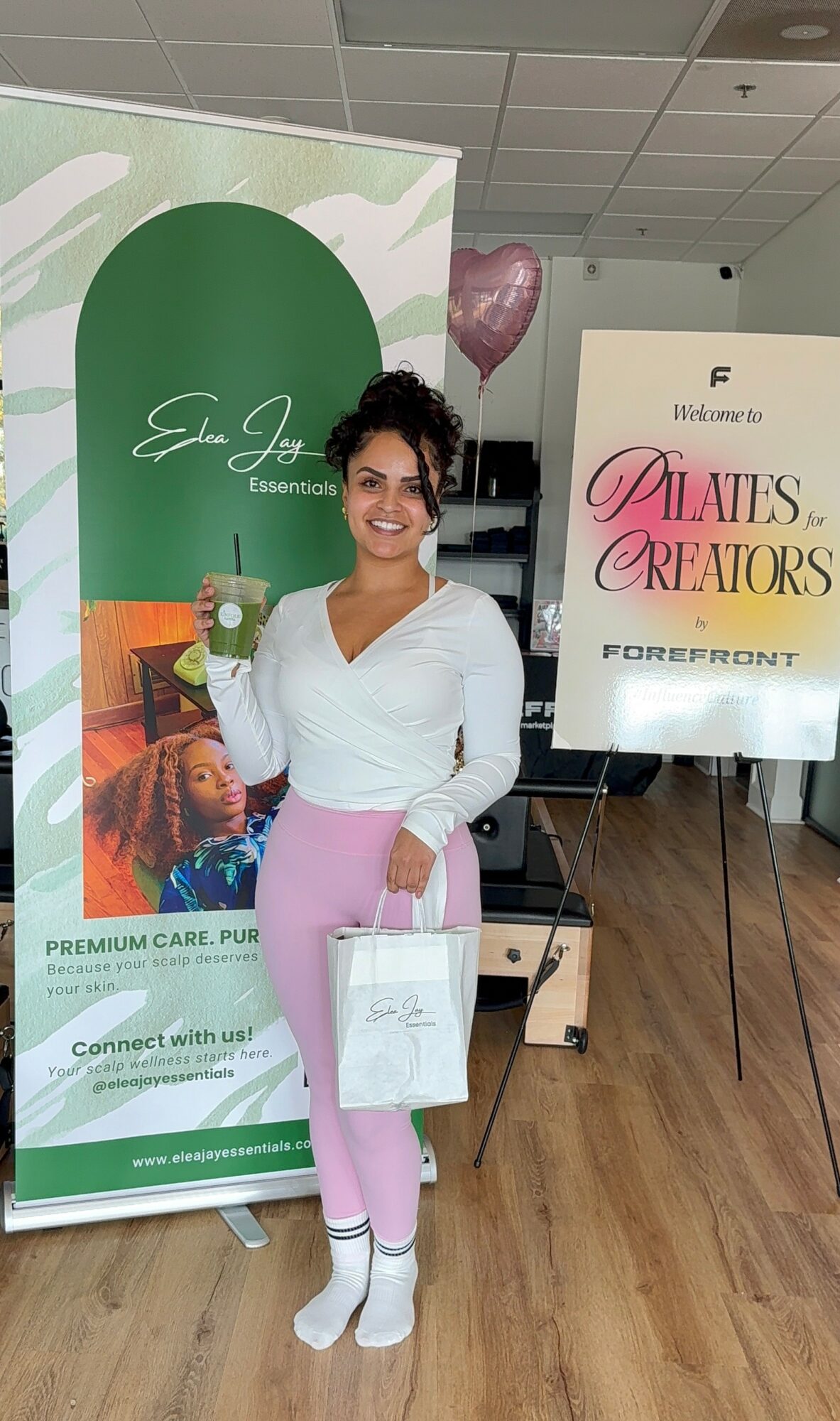 Smiling woman holding a gift bag standing next to a green and white banner and a sign with pink and black text in an indoor setting.