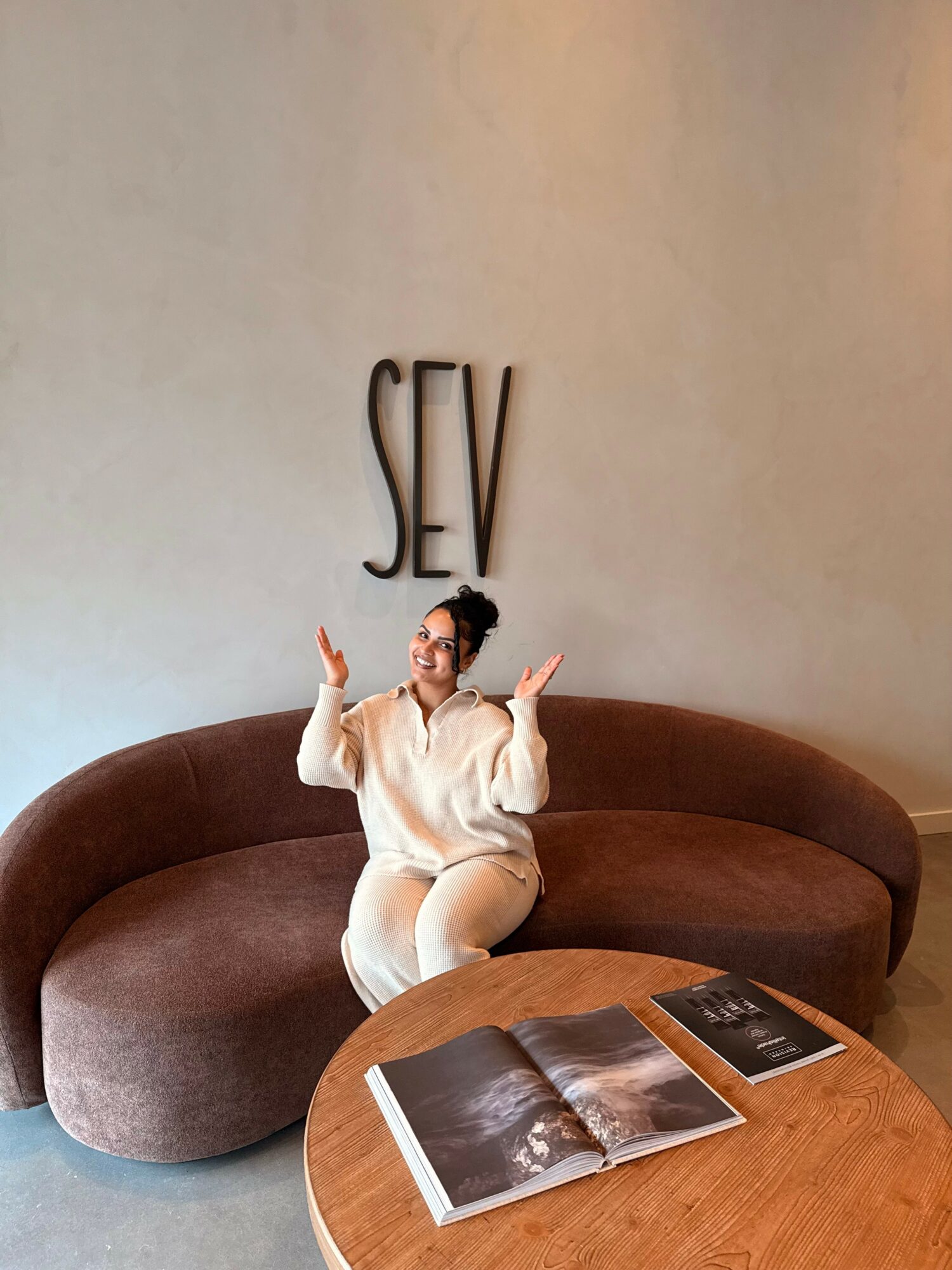 Person sitting on a brown curved sofa with arms raised, in front of a wall with large 'SEV' letters, and a wooden table with open magazines.