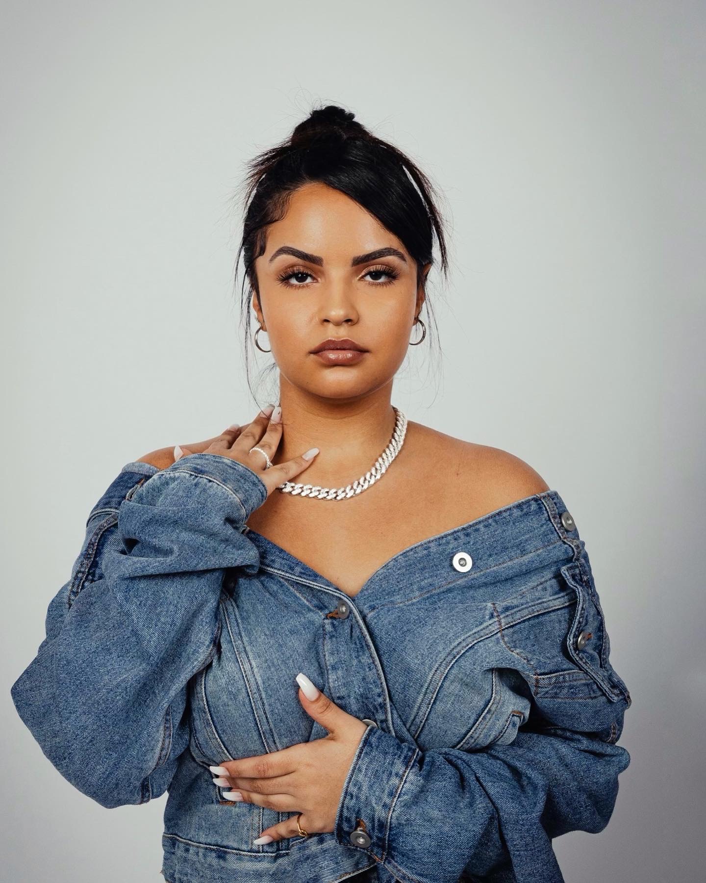 Woman with dark hair in a bun, wearing a denim jacket off her shoulders, jewelry, and makeup, looking at the camera.