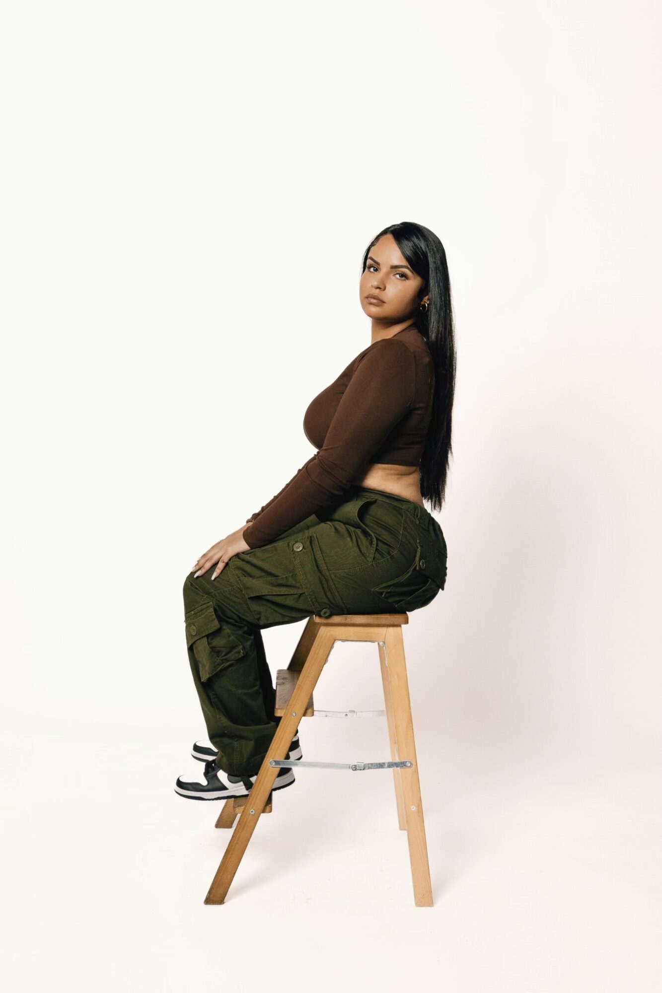 Woman with long dark hair sitting sideways on a wooden stool, wearing a brown top and green pants, against a plain white background.