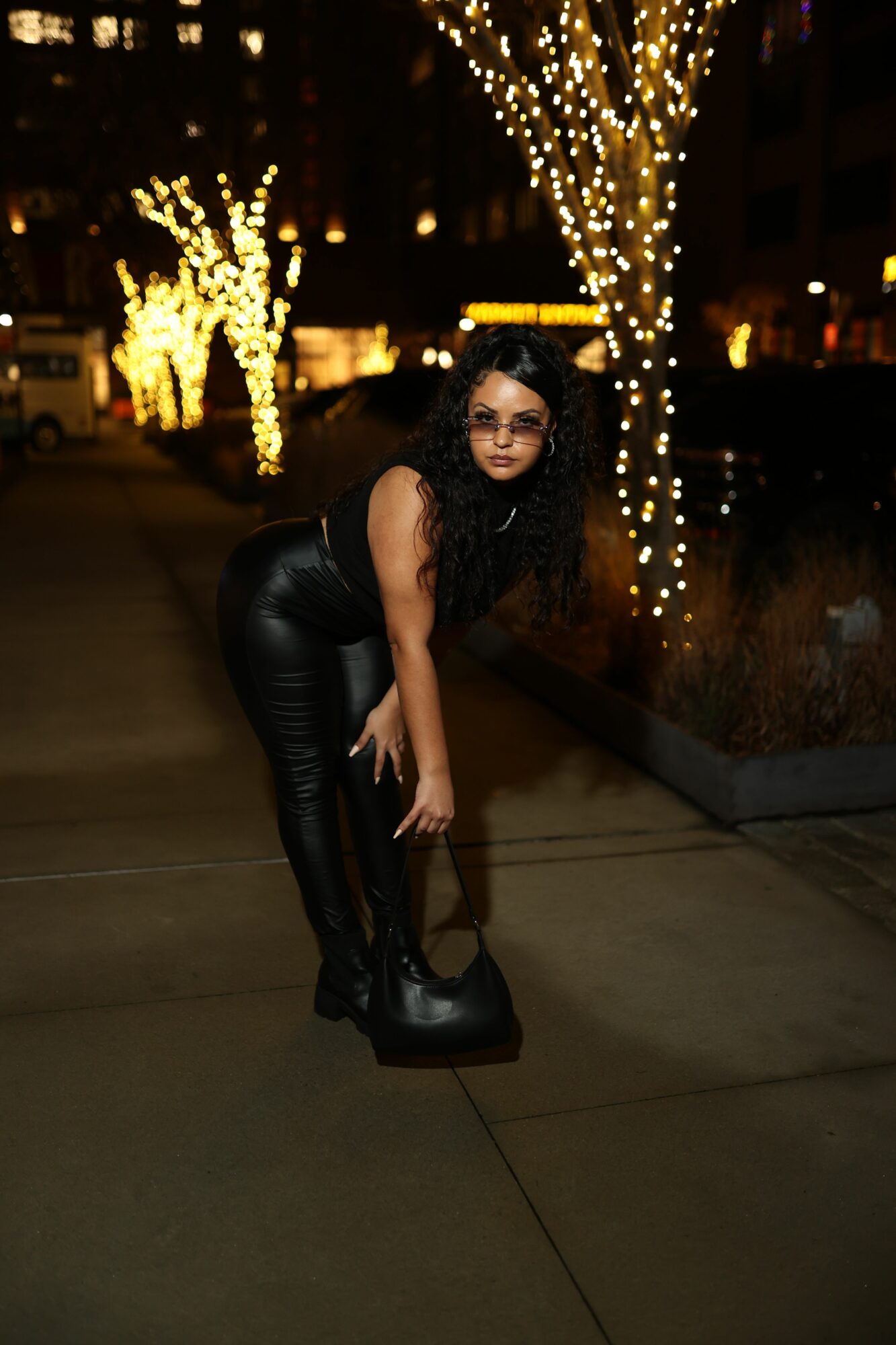 Woman with black hair in a black leather outfit standing on a sidewalk at night, illuminated by string lights and city lights.