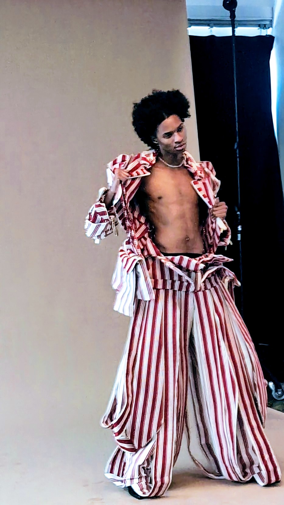 Person with curly hair in striped pants and jacket, shirtless, standing against a plain background.