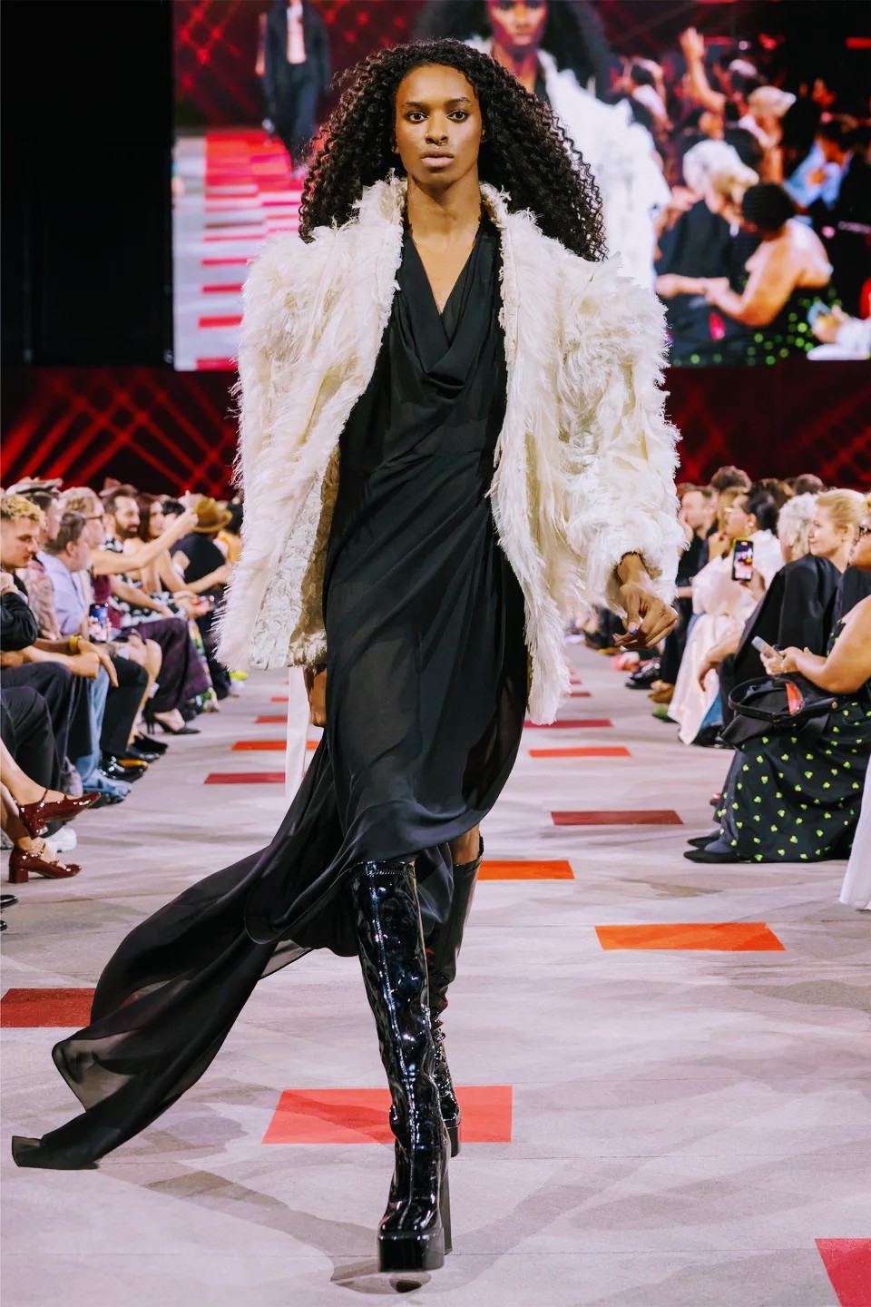 Model walking down runway in black dress, white fur coat, and high-heeled boots, audience seated on both sides, large screen behind.